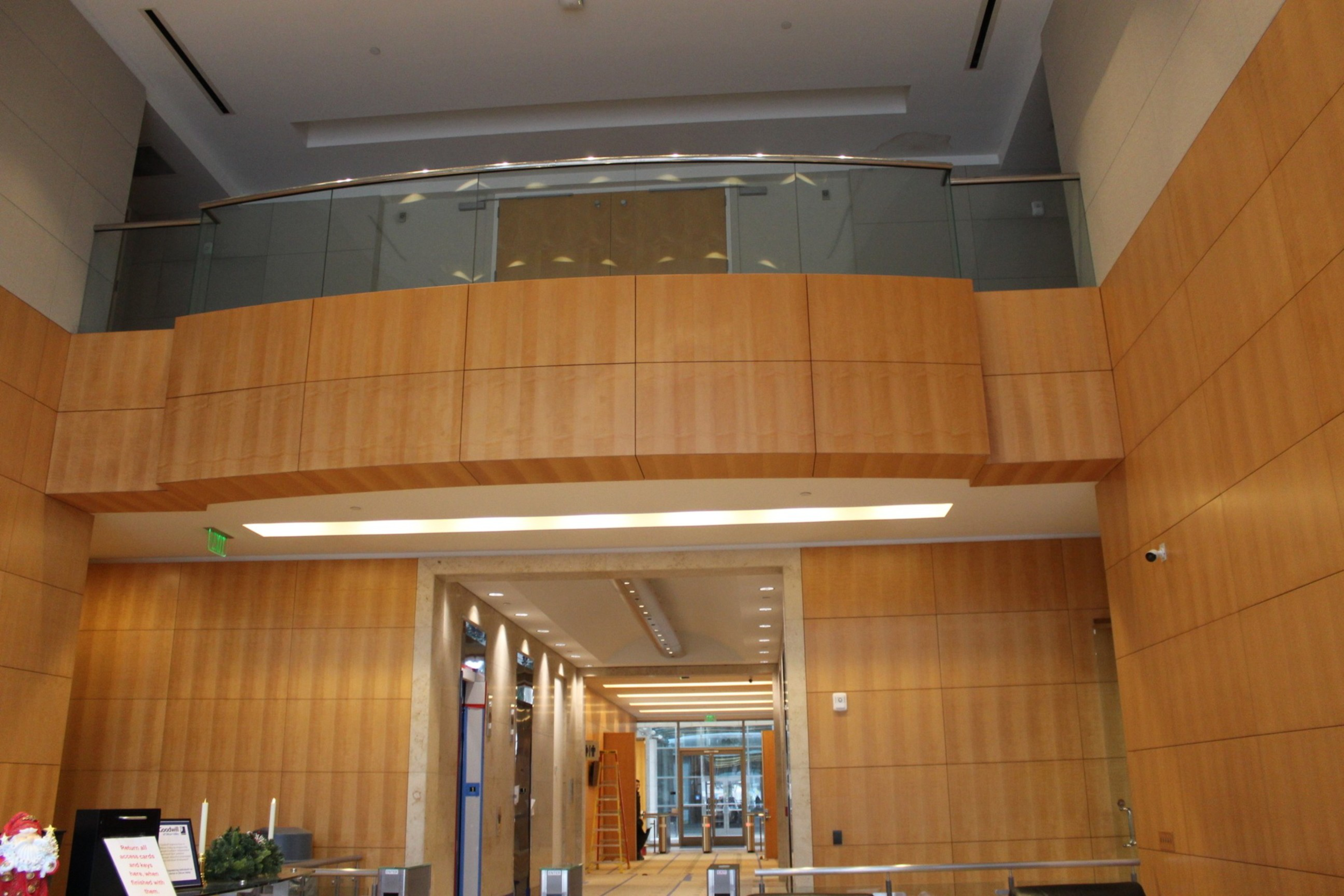 A modern indoor hallway with wood-paneled walls, a glass balcony above, and ceiling lights leads to a bright entrance with metal turnstiles.