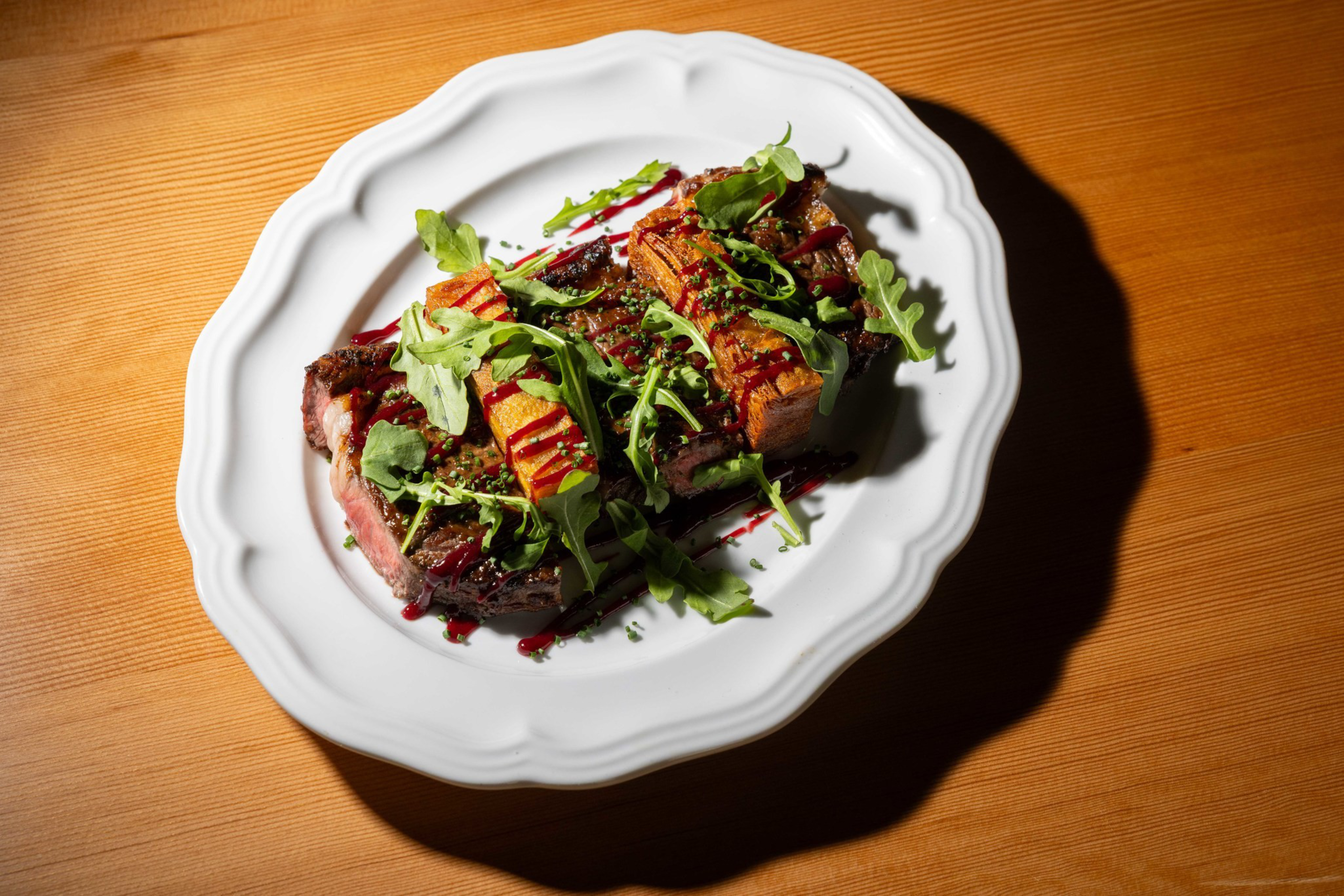 Slices of steak topped with arugula, crispy rectangular potato pieces, green herbs, and drizzled with a dark red sauce on a white plate.