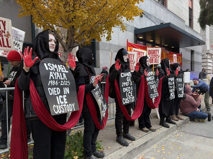 A group of people in black robes and red sashes hold signs naming individuals who died in ICE custody during a protest outside a building.