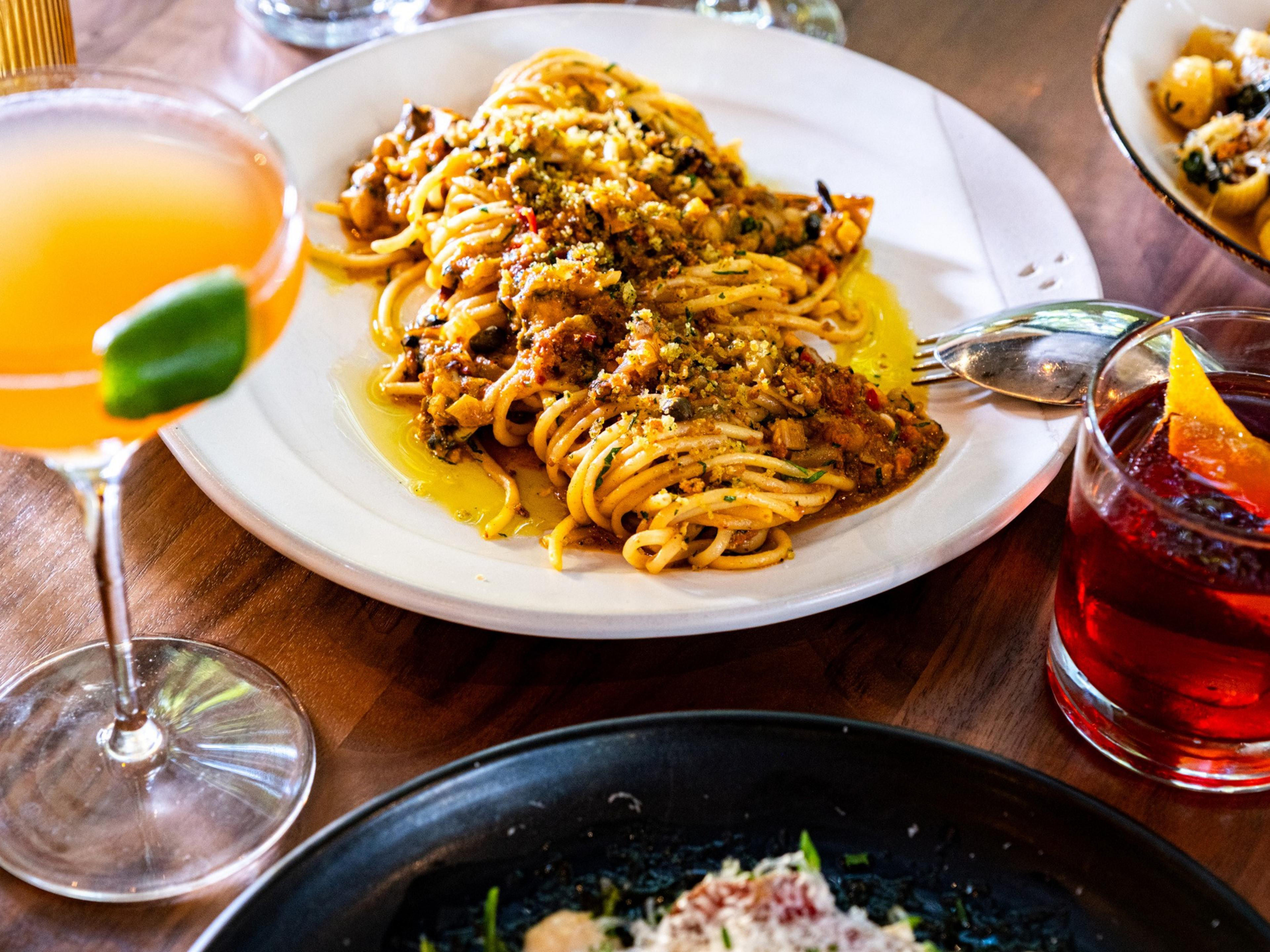 A plate of spaghetti with meat sauce and grated cheese is surrounded by two cocktails, one orange with a lime twist and one dark with an orange slice.