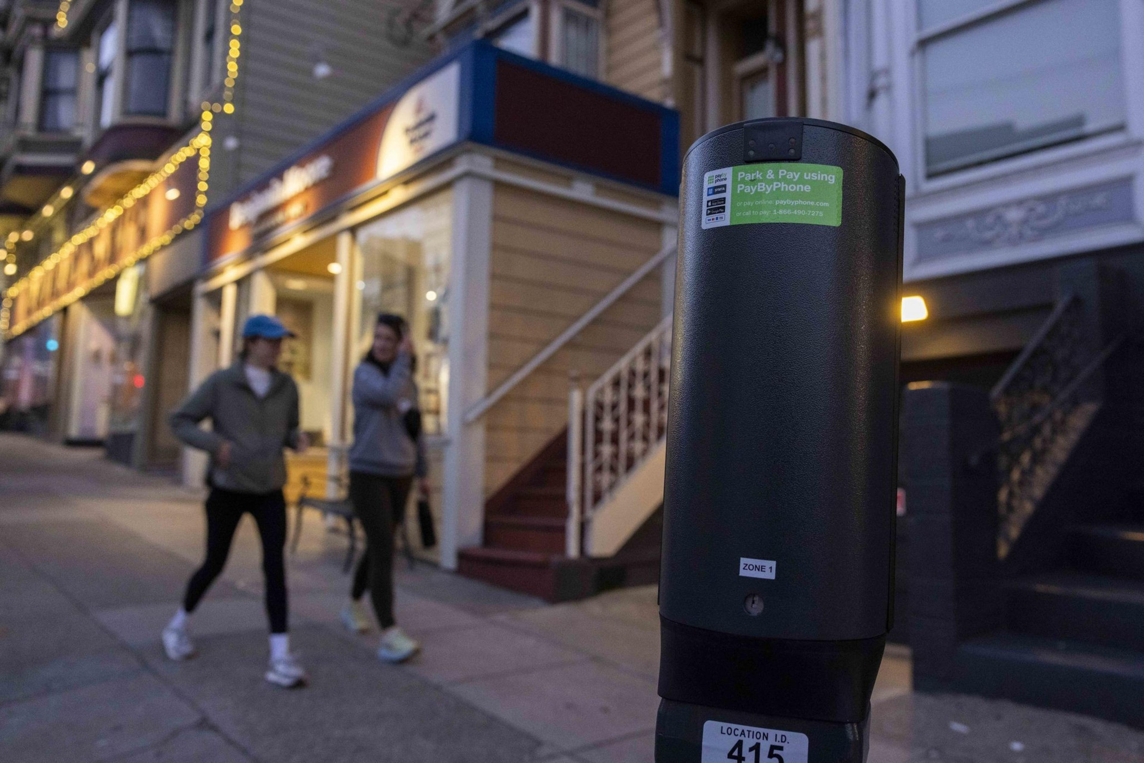 A parking meter is in the foreground with instructions for PayByPhone. Two women walk on the sidewalk past a store with string lights and a yellow faÃ§ade.