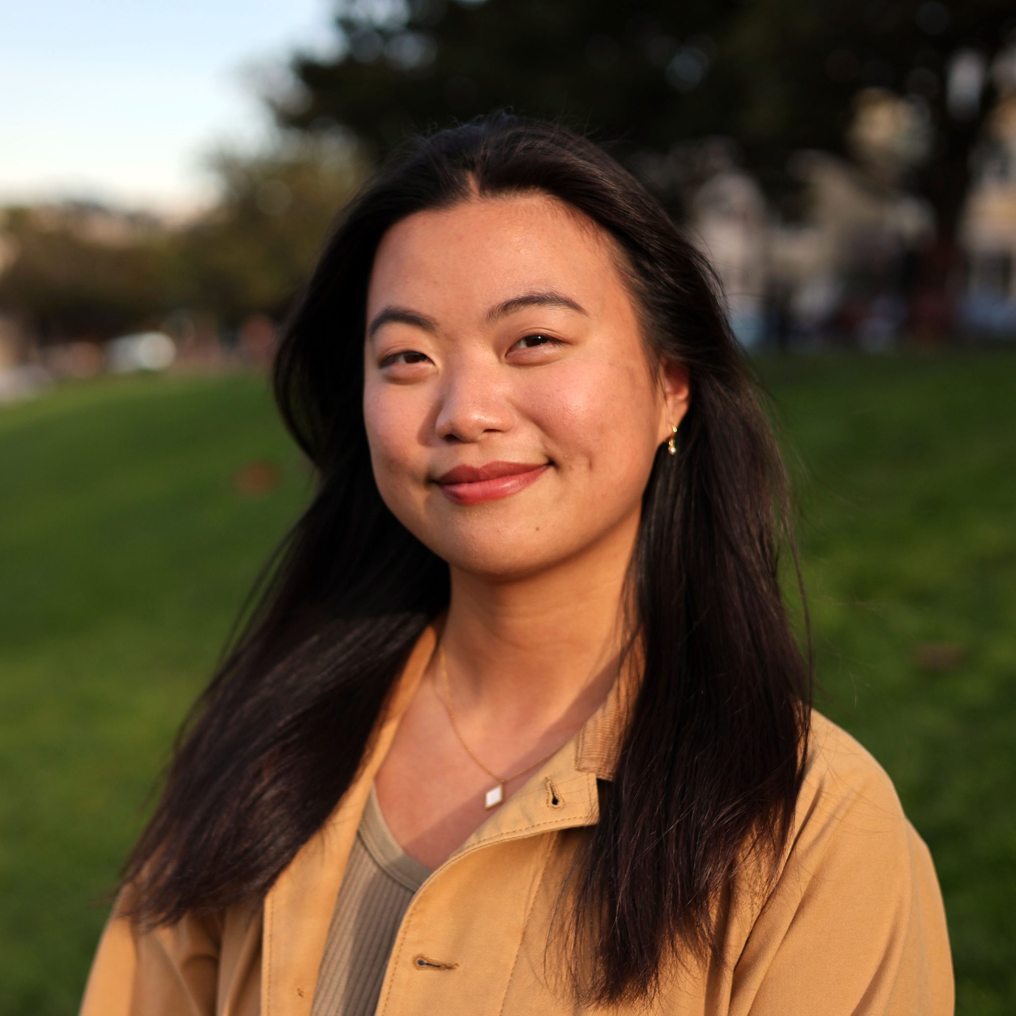A person with long black hair smiles softly, standing outdoors in a park. They are wearing a tan jacket and a necklace. The background is grassy with trees slightly blurred.