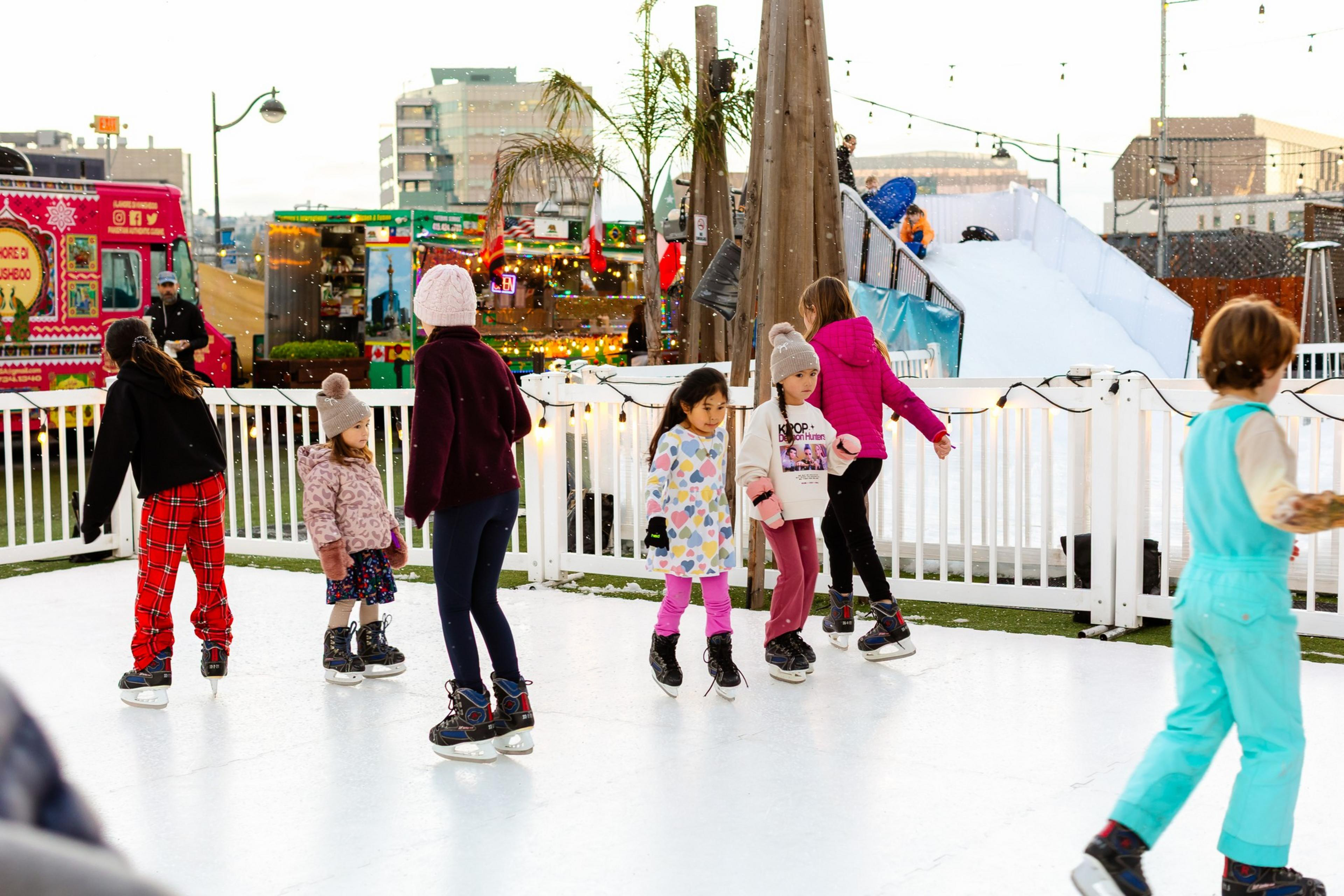 Children wearing winter clothes skate on an outdoor ice rink with a white fence, festive lights, and food trucks in the background.