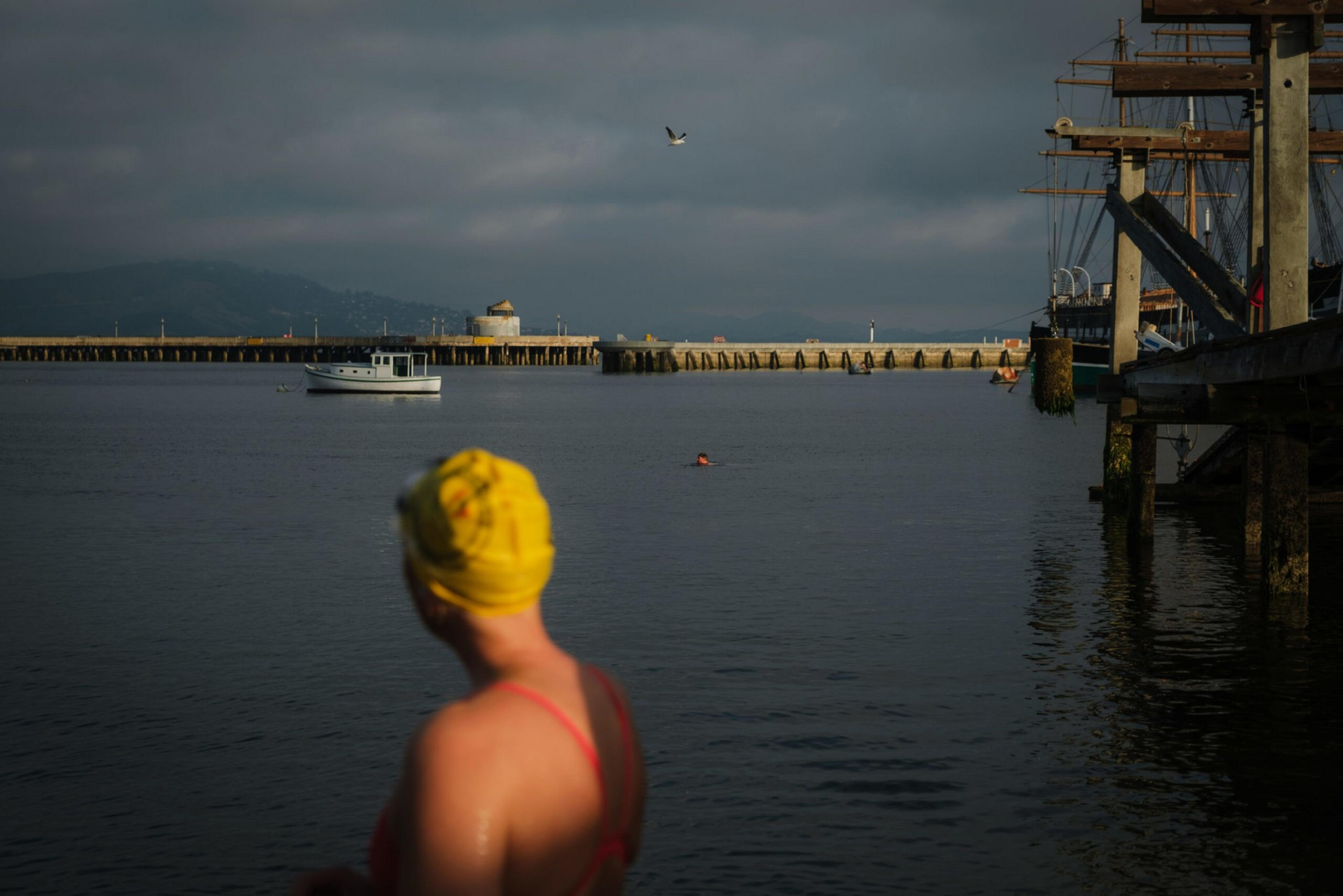 A swimmer wearing a yellow swim cap looks out over calm water towards a boat, a pier, and a distant shoreline under a cloudy sky.