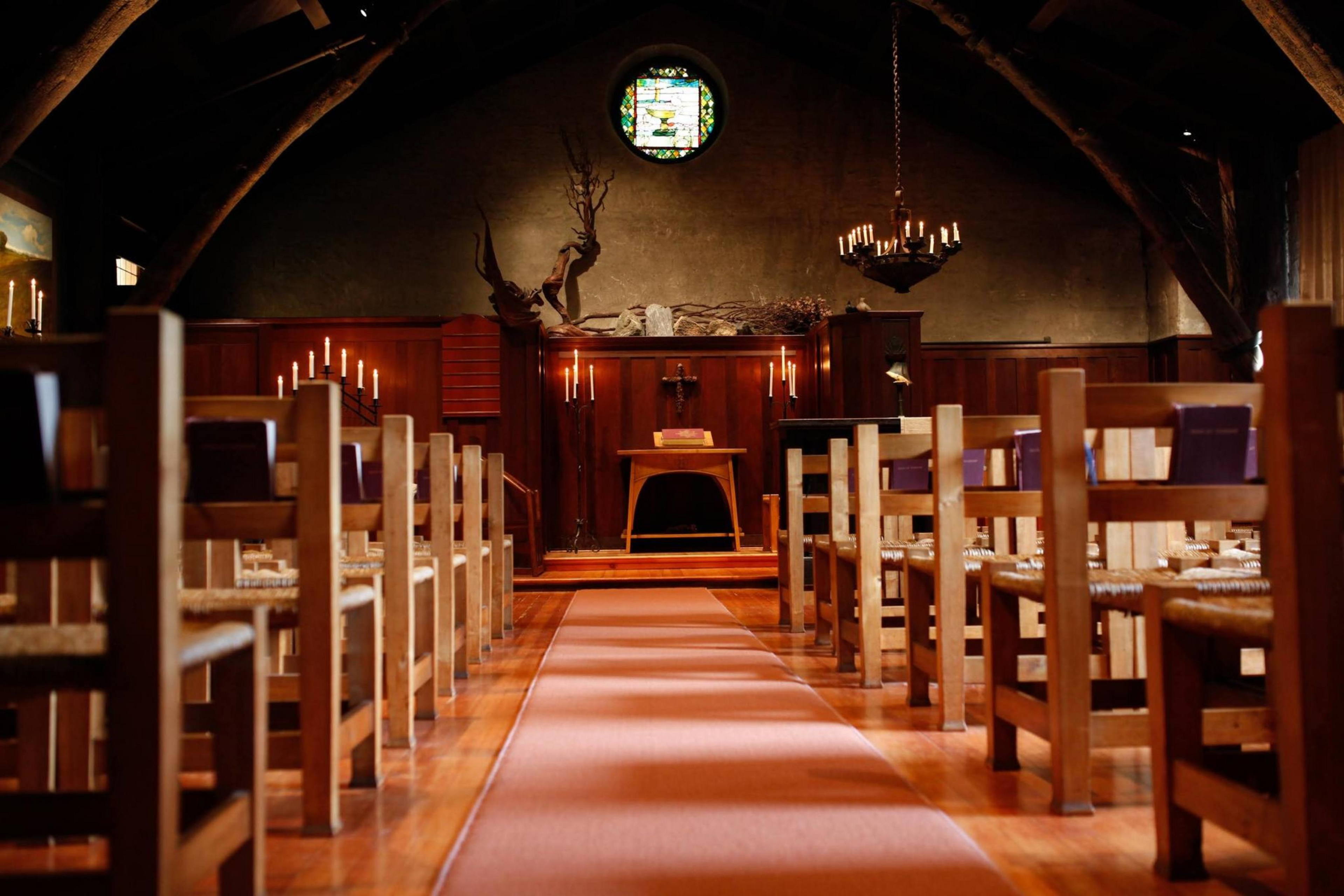 A narrow aisle runs between wooden pews facing a simple altar, with dim lighting, candles, a stained-glass window, and antlers mounted above the altar.