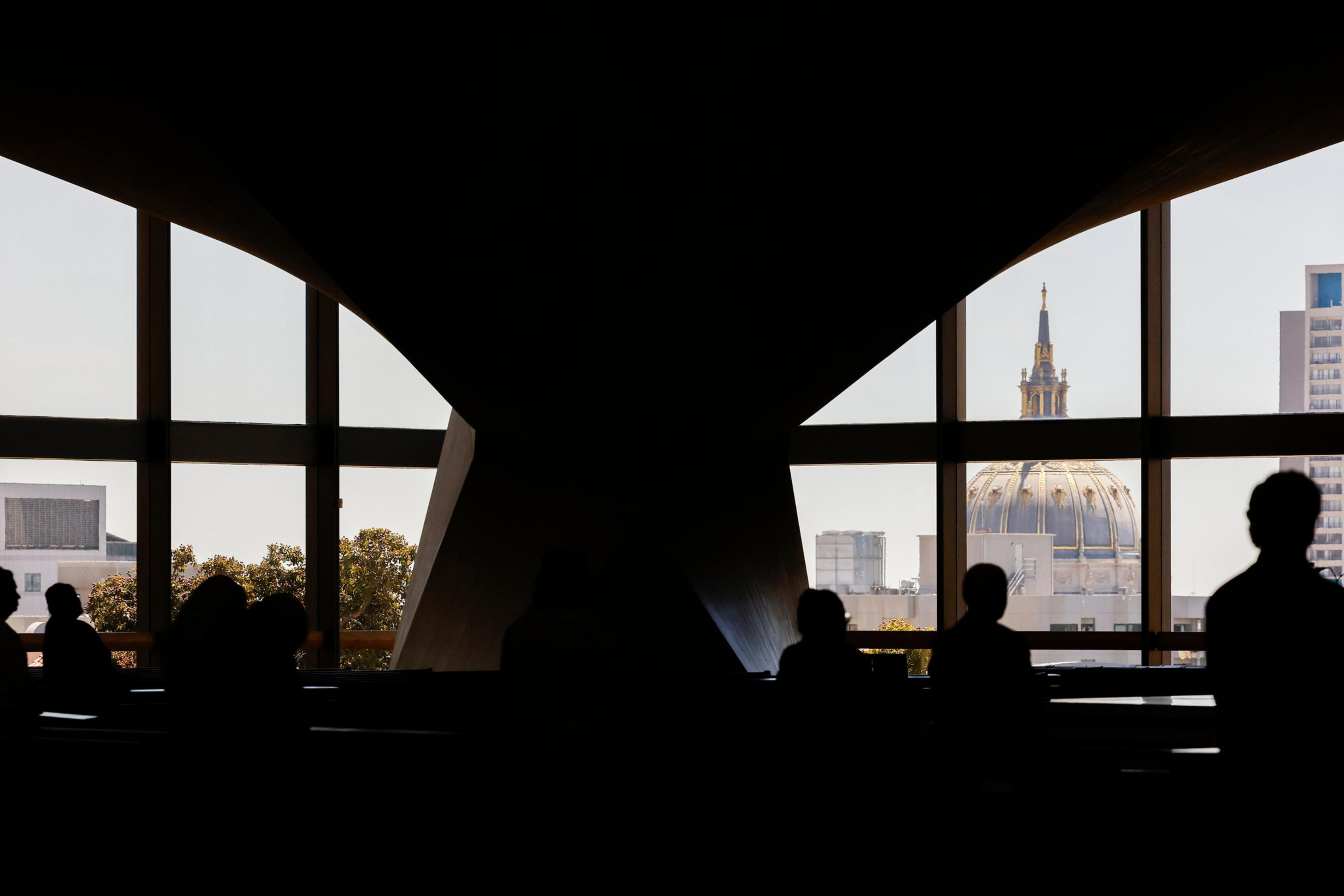 Silhouetted people sit near large windows framing a cityscape with trees, buildings, and a prominent domed structure topped with a spire.