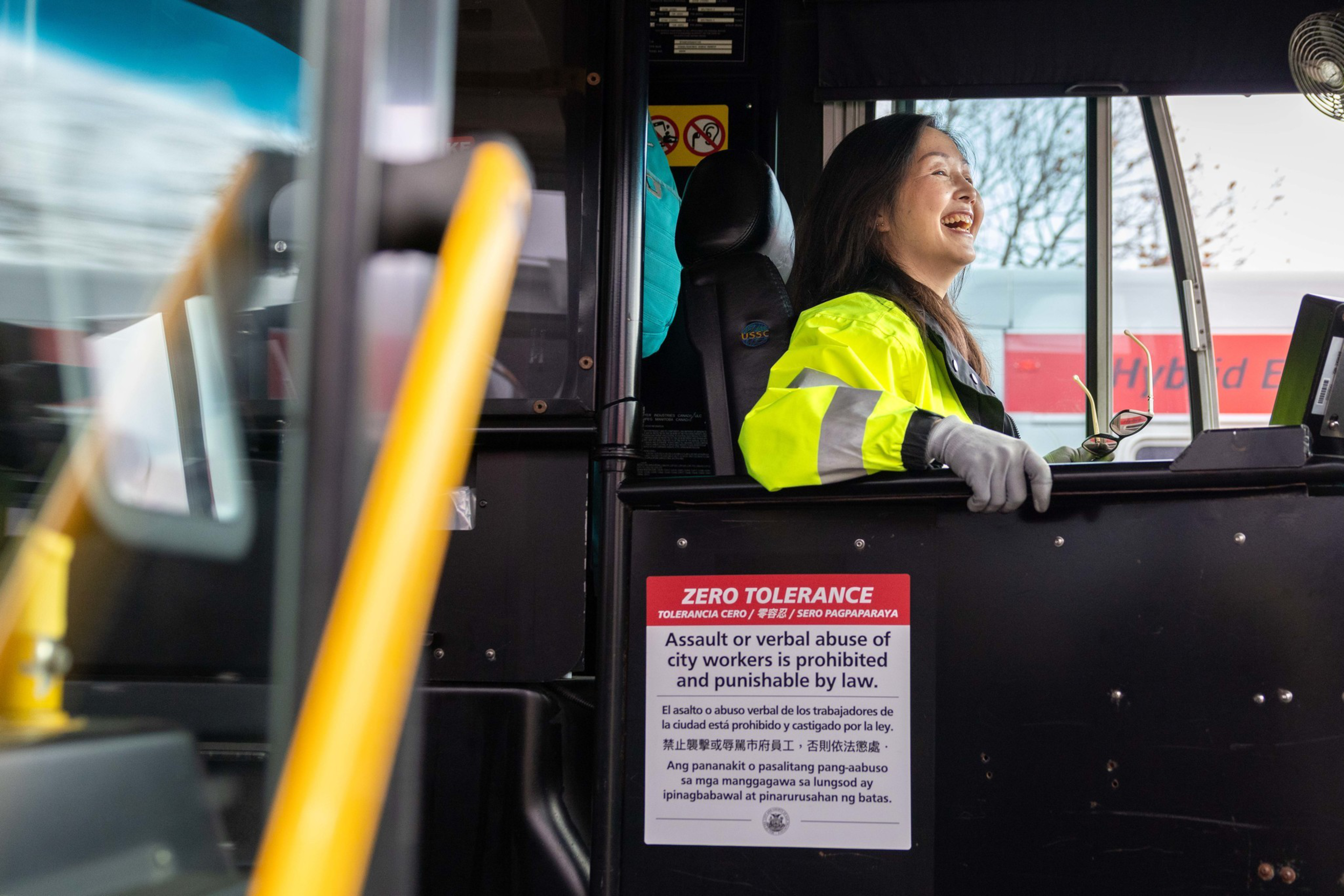 A woman in a bright yellow safety jacket and gloves smiles while sitting at the driver’s seat of a city bus, with a “Zero Tolerance” sign visible.