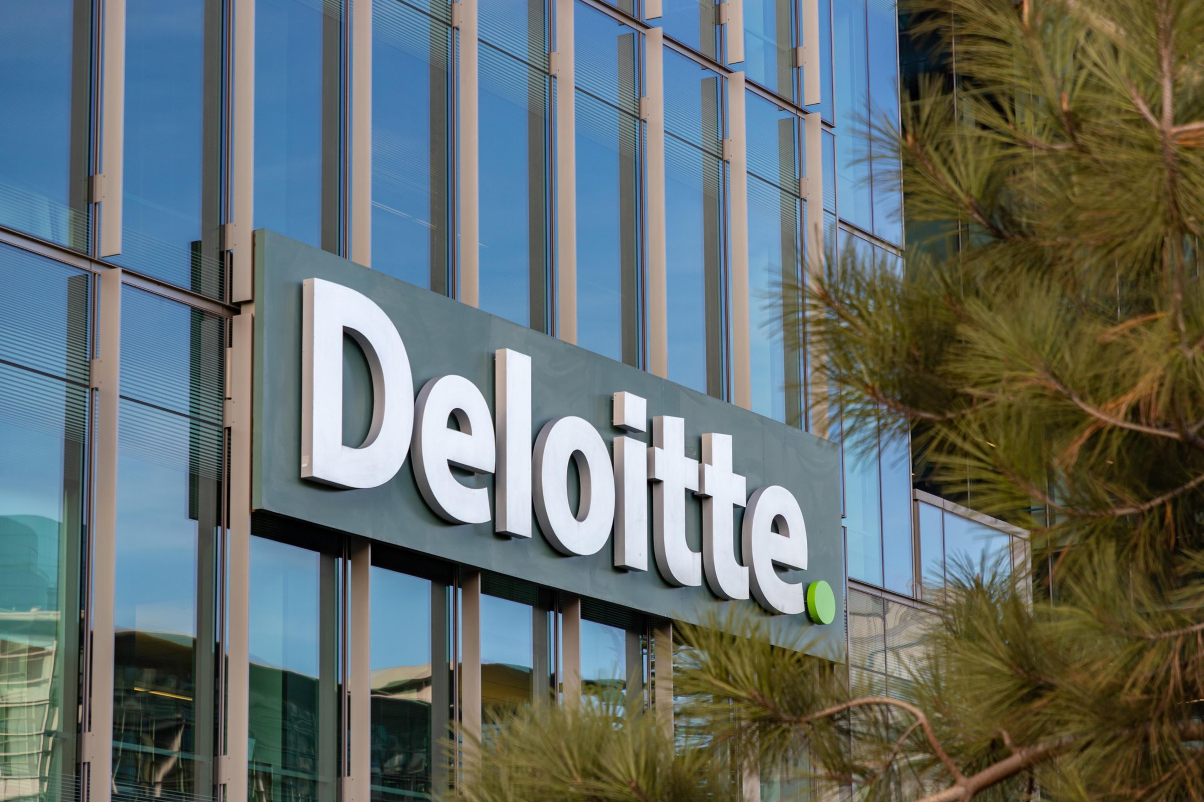 The image shows a modern glass building with the “Deloitte” logo prominently displayed in white letters. Pine branches are partially visible on the right.