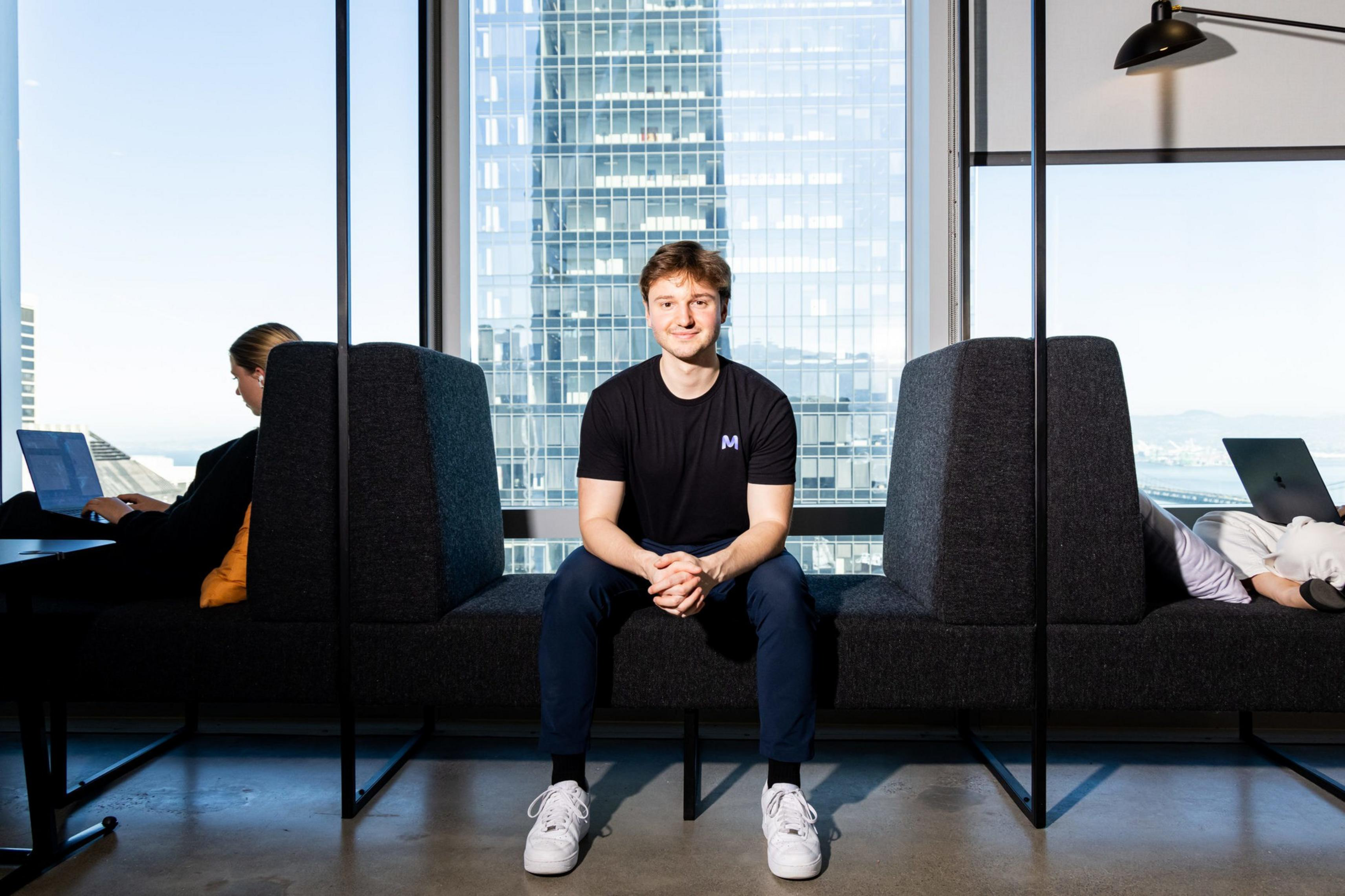 A young man in a black shirt and white sneakers sits on a dark bench in front of a large window with a cityscape behind him. Two people work on laptops at either side.