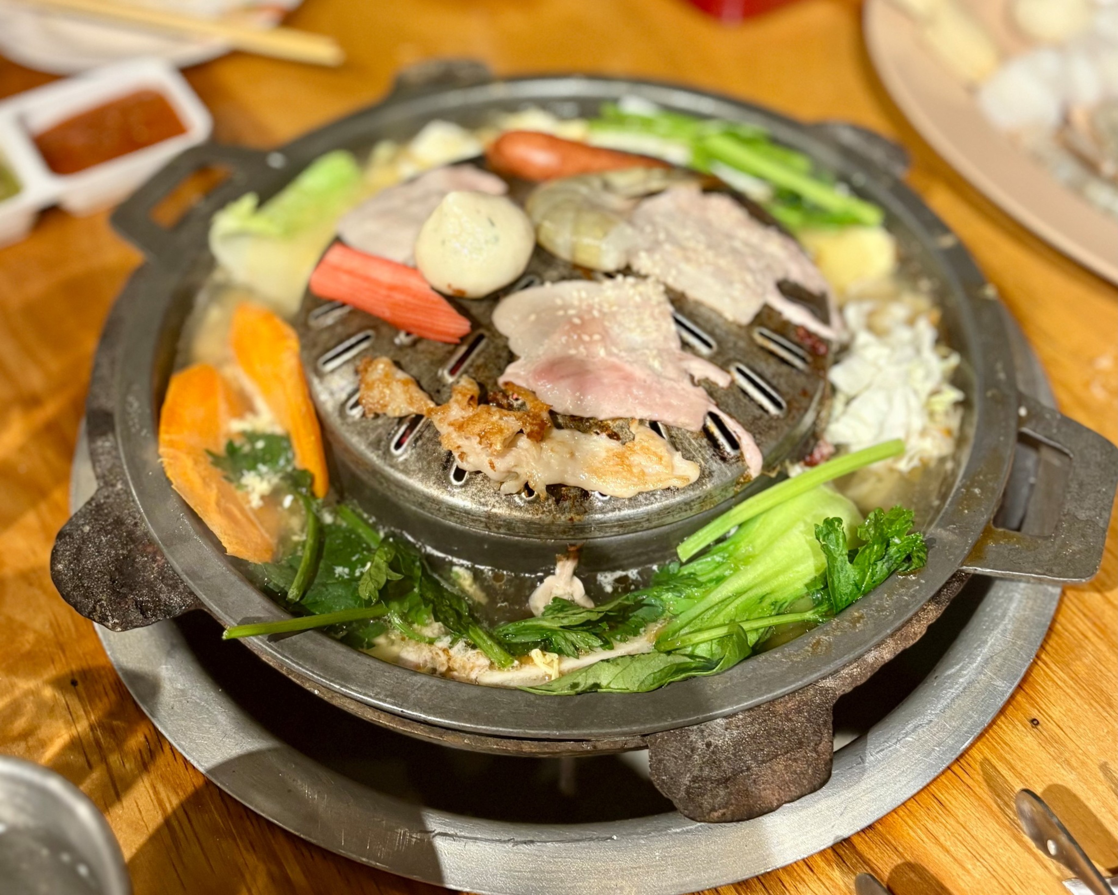A Korean BBQ grill with slices of meat, seafood, vegetables, and a broth-filled outer ring containing greens and egg cooking.