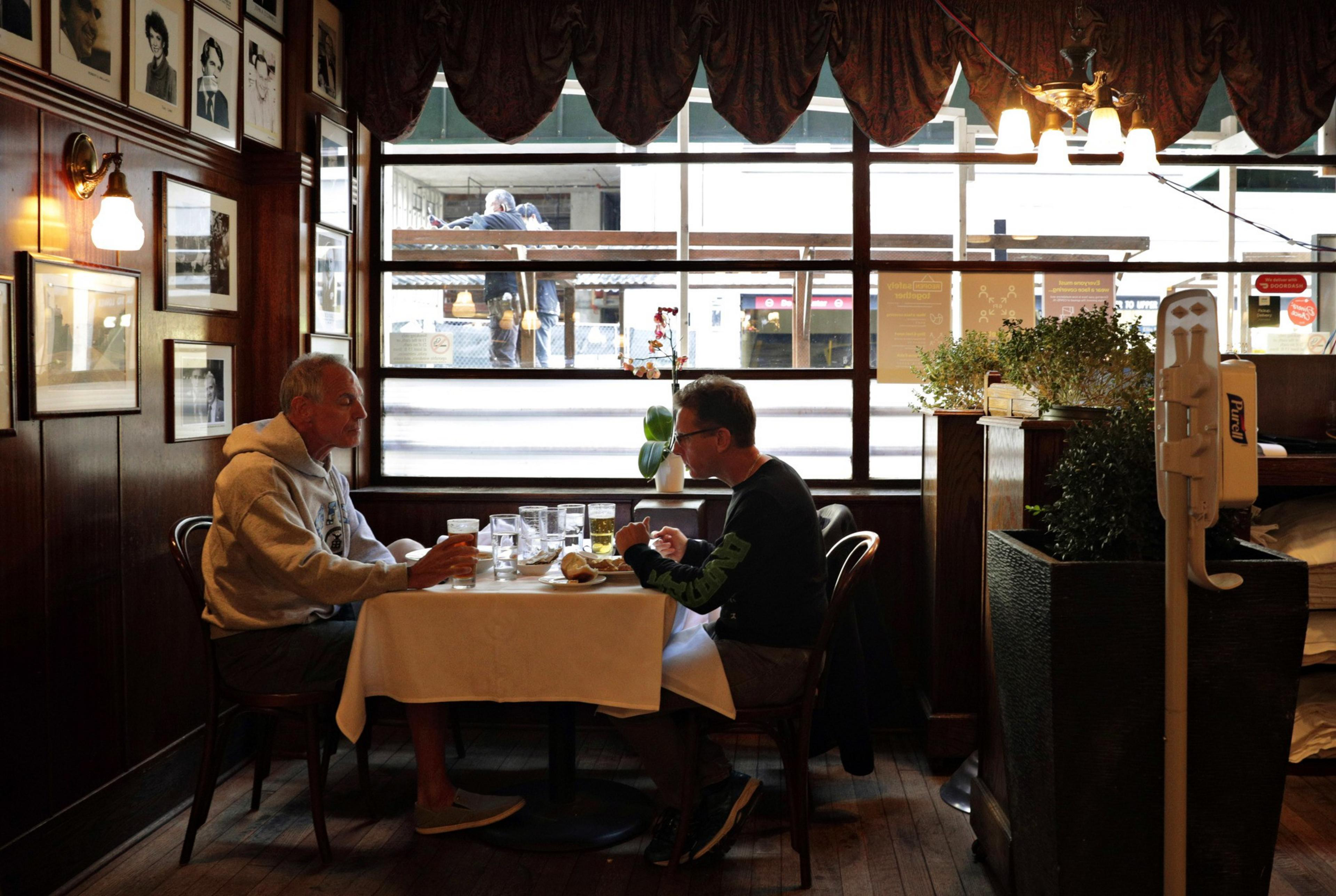Two men sit at a table in a cozy restaurant, surrounded by framed photos on wood-panelled walls. Large windows reveal a street scene outside.