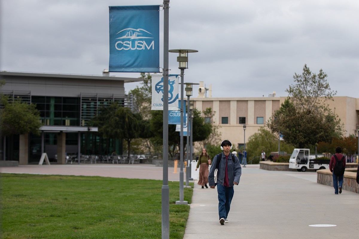A person wearing headphones walks along a pathway near lamp posts banners with the blue Cal State San Marcos logo on them.