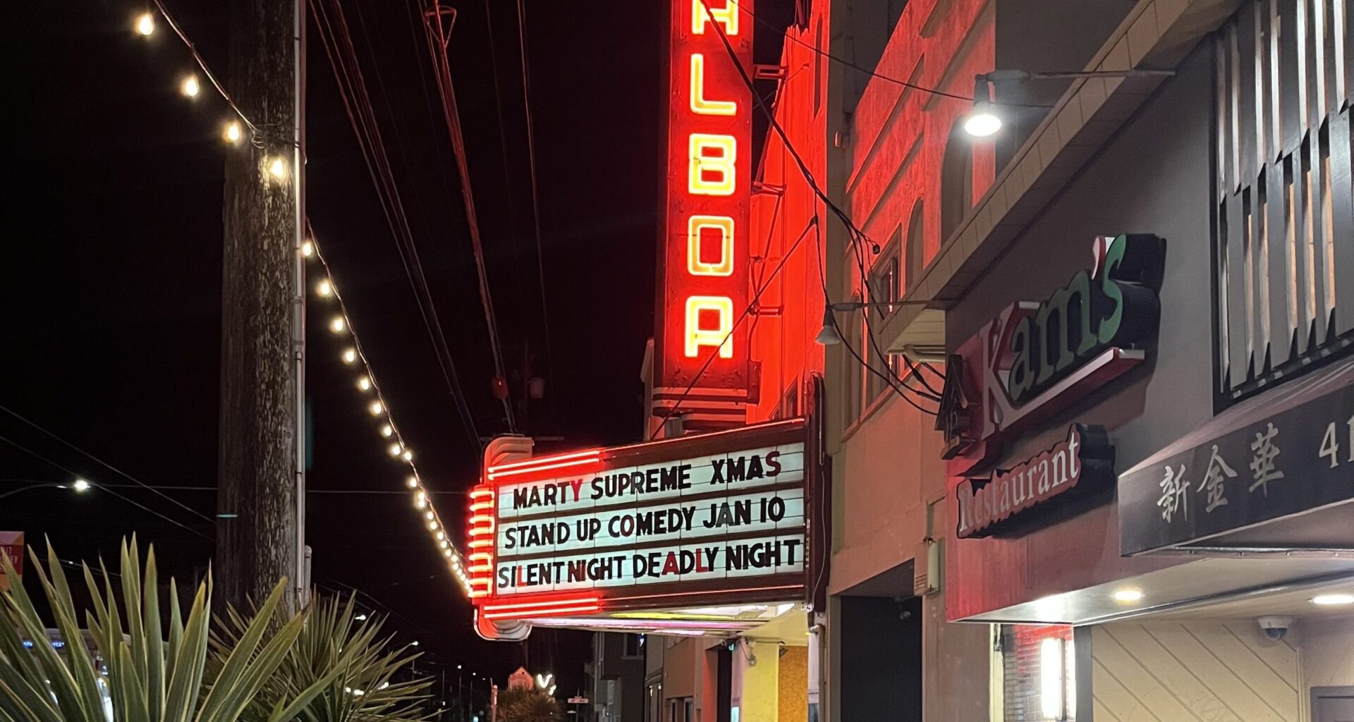 A sidewalk view of the Balboa theater at night with a red vertical neon sign and marquee listing upcoming events, next to Kam