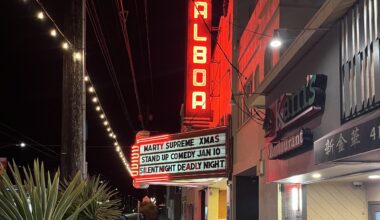 A sidewalk view of the Balboa theater at night with a red vertical neon sign and marquee listing upcoming events, next to Kam