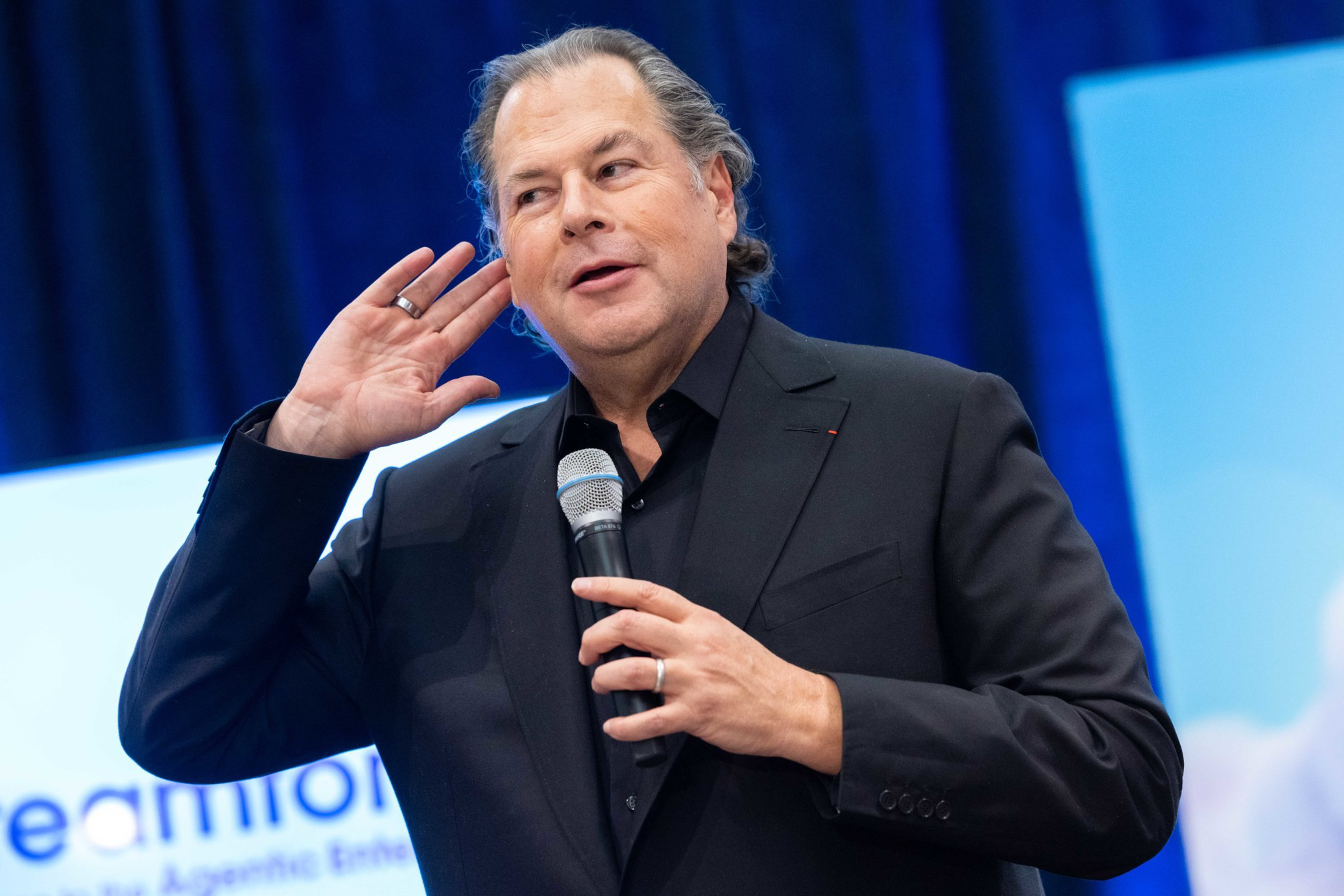 A man in a black suit holds a microphone in one hand and cups his ear with the other, as if listening intently.