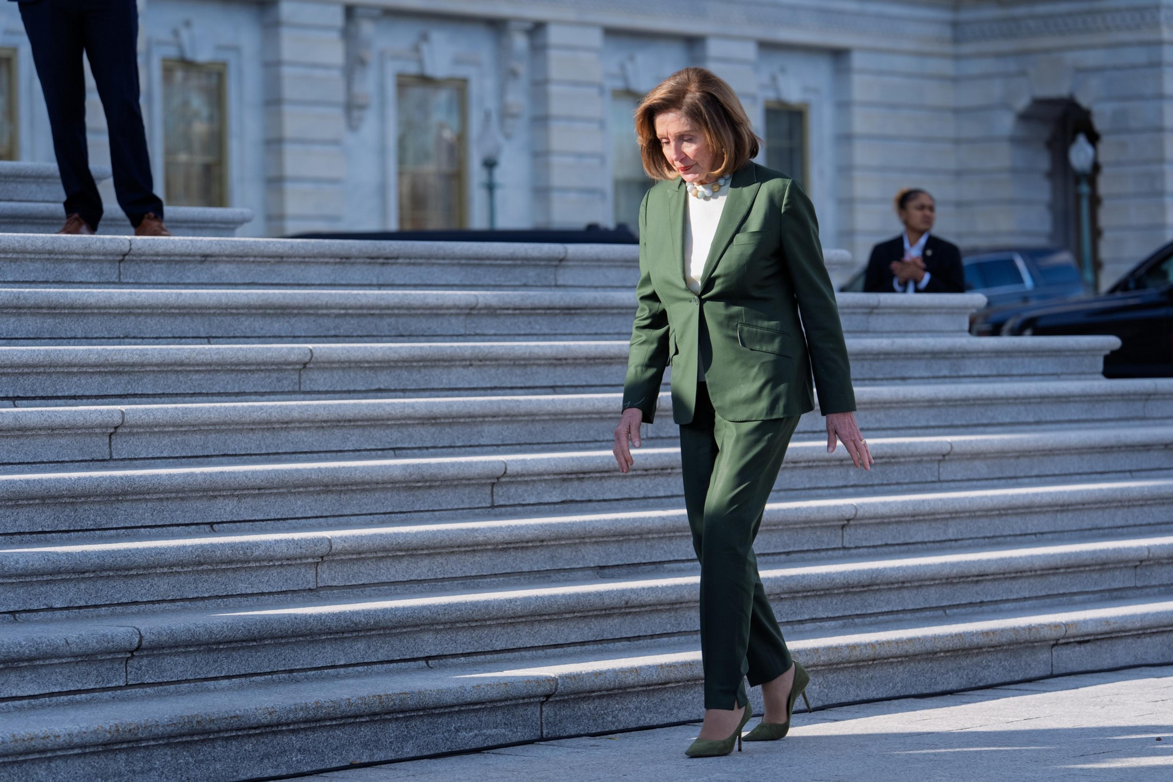 A woman in a green pantsuit and heels walks down stone steps outside a large government building in daylight.