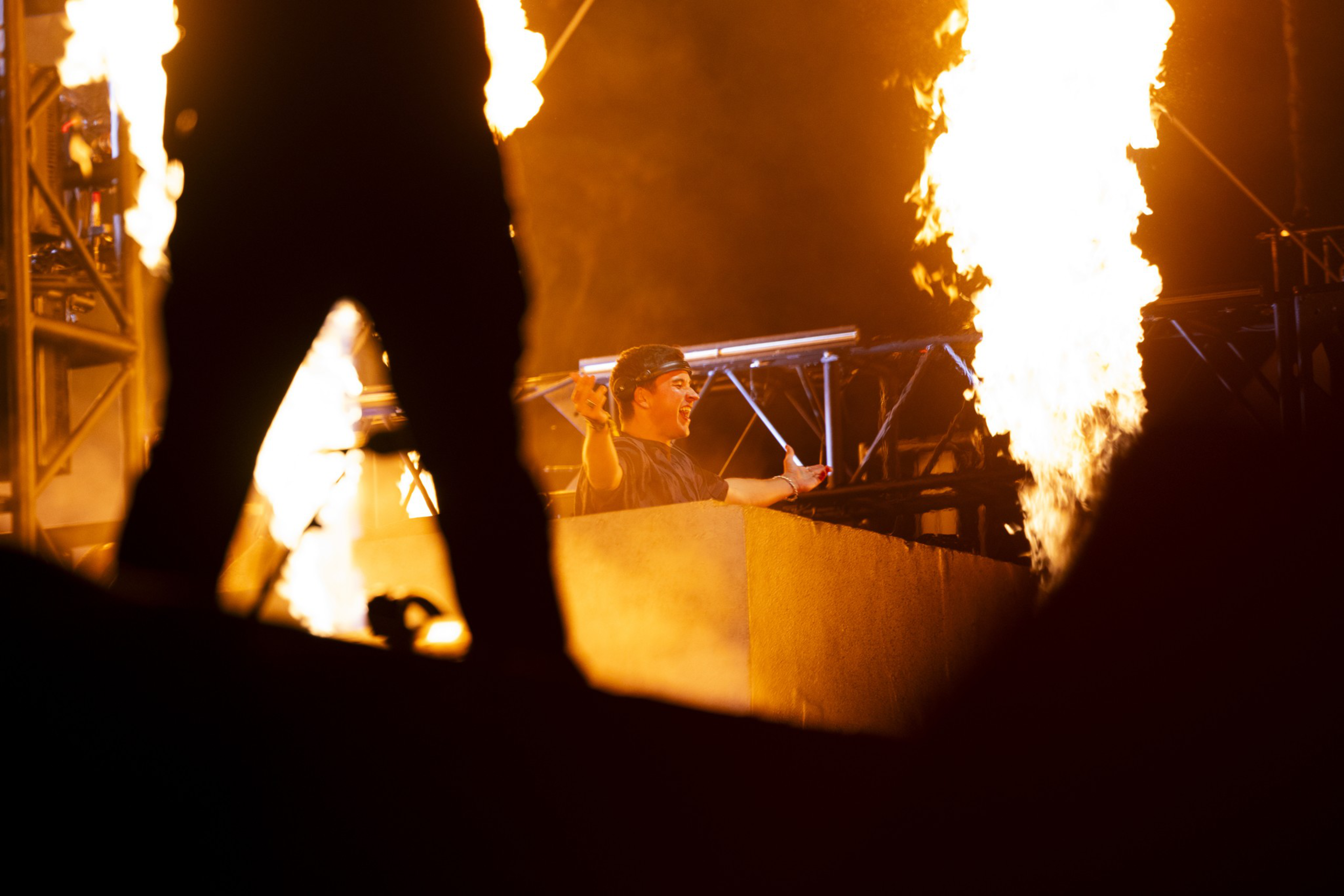 A DJ performs on stage amid large vertical flames, while a silhouetted figure stands in the foreground, partially blocking the view.