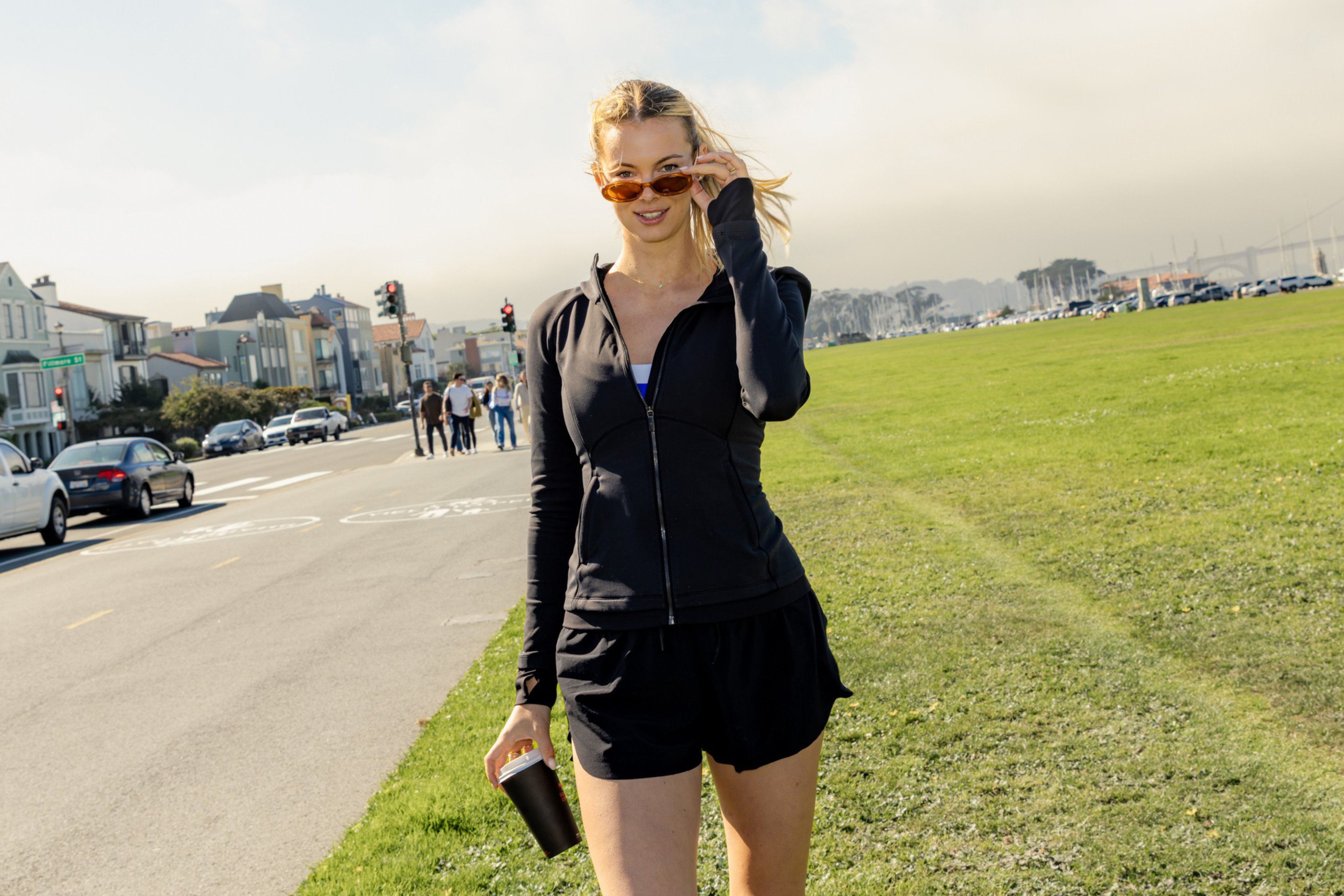 A woman in black athletic wear holds a black tumbler, adjusting her sunglasses, standing on grass beside a street with cars and people.