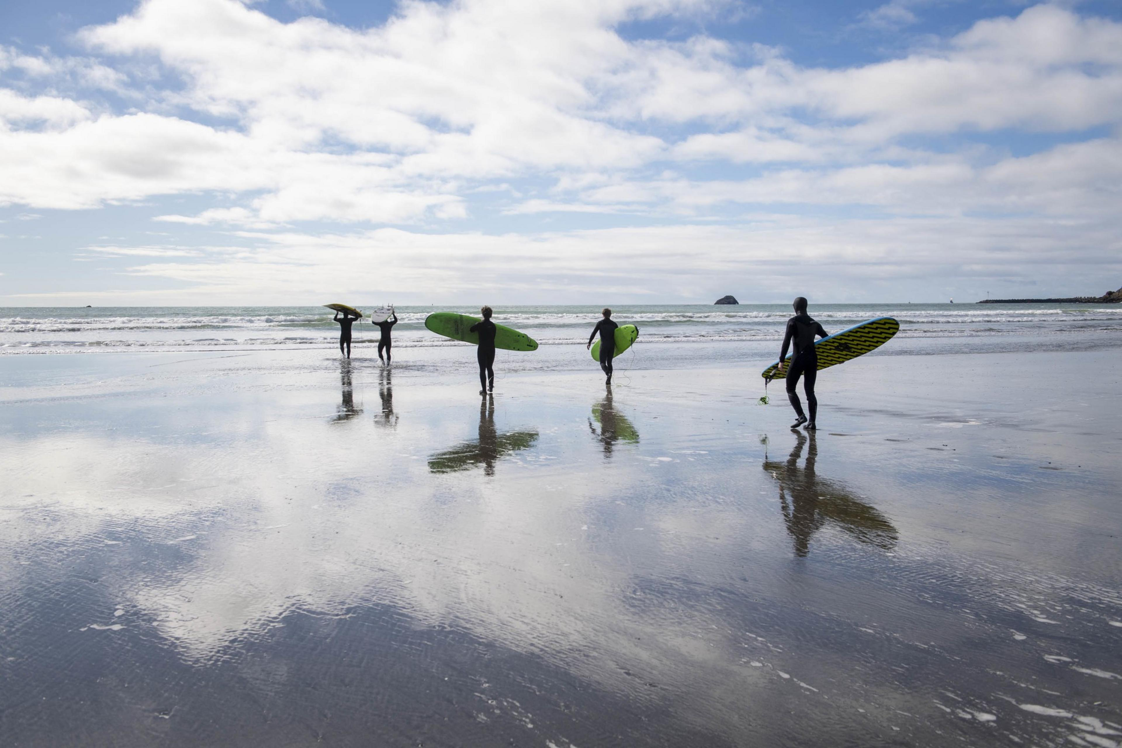 Five surfers in wetsuits walk towards the ocean, carrying colorful surfboards. The sky is partly cloudy, and the wet sand reflects the clouds above.