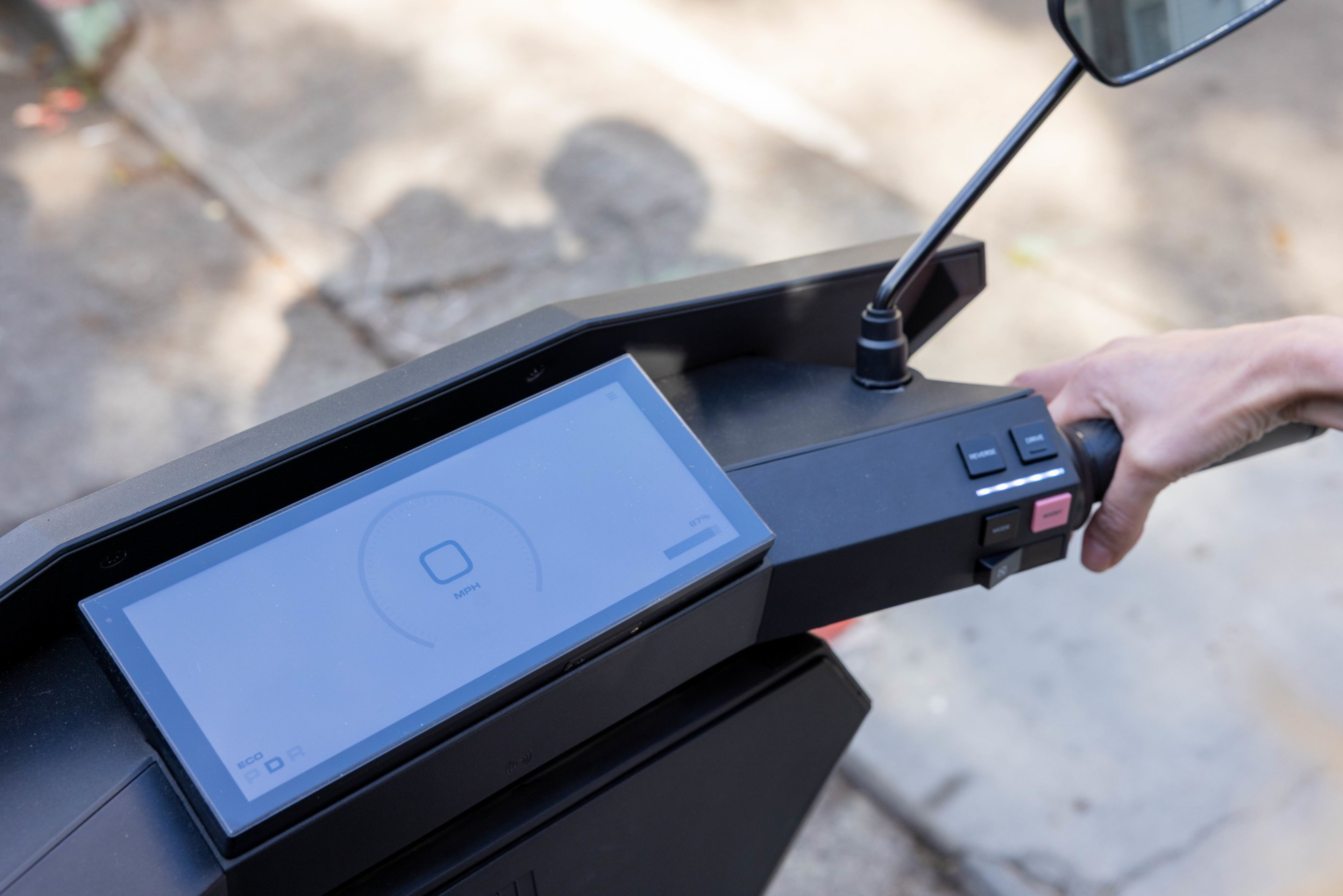 A close-up of a scooter handlebar with a digital display showing a circular loading icon and buttons near the right hand gripping the handle.