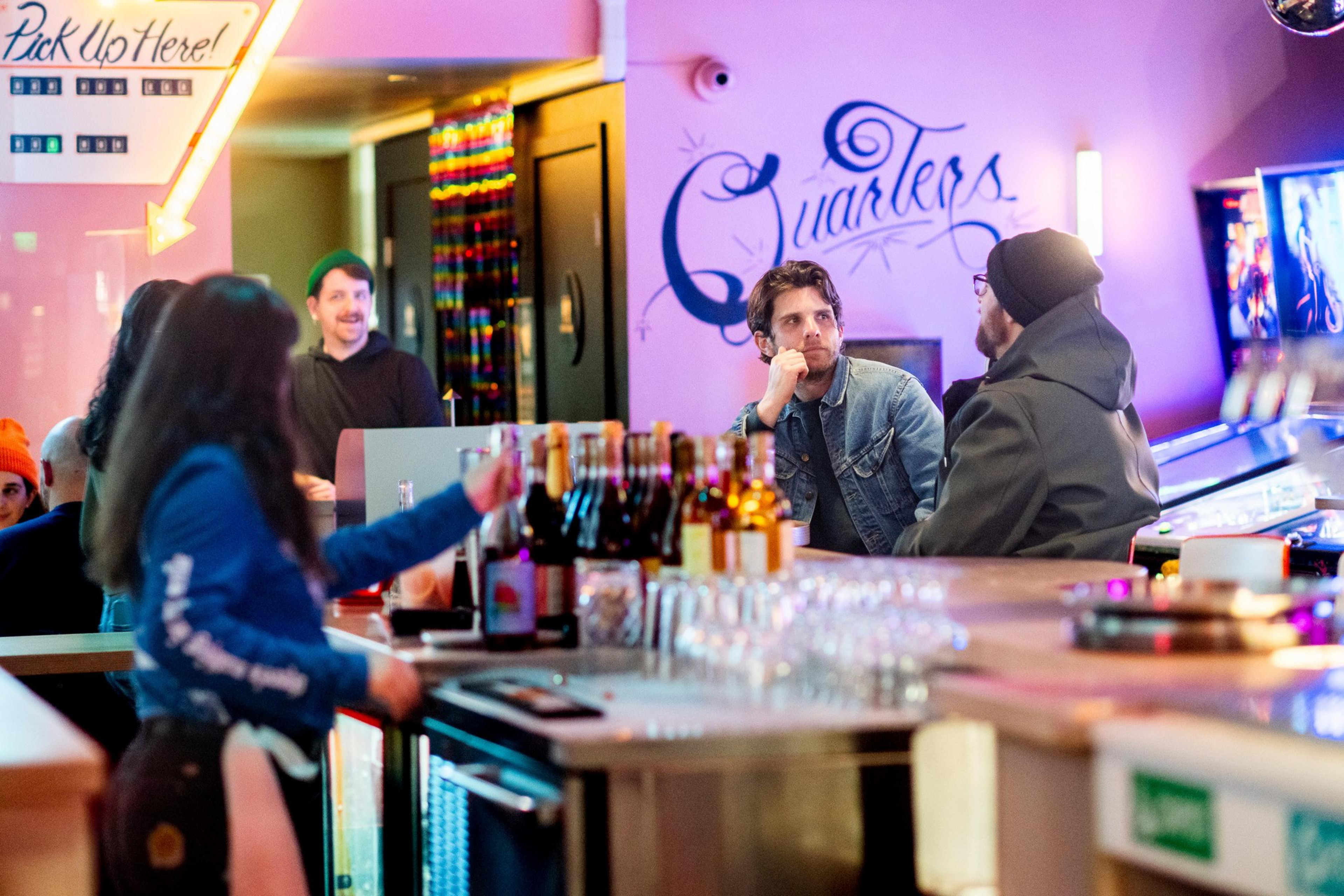 A lively bar scene with people chatting. A person in a denim jacket sits at the counter, while others are gathered nearby. Bottles and glasses line the bar.