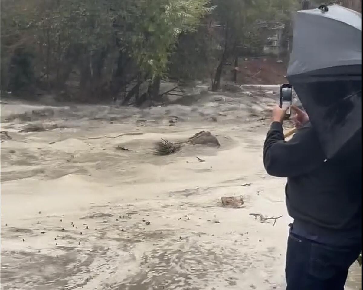 A person in black with an umbrella stands near floodwaters 