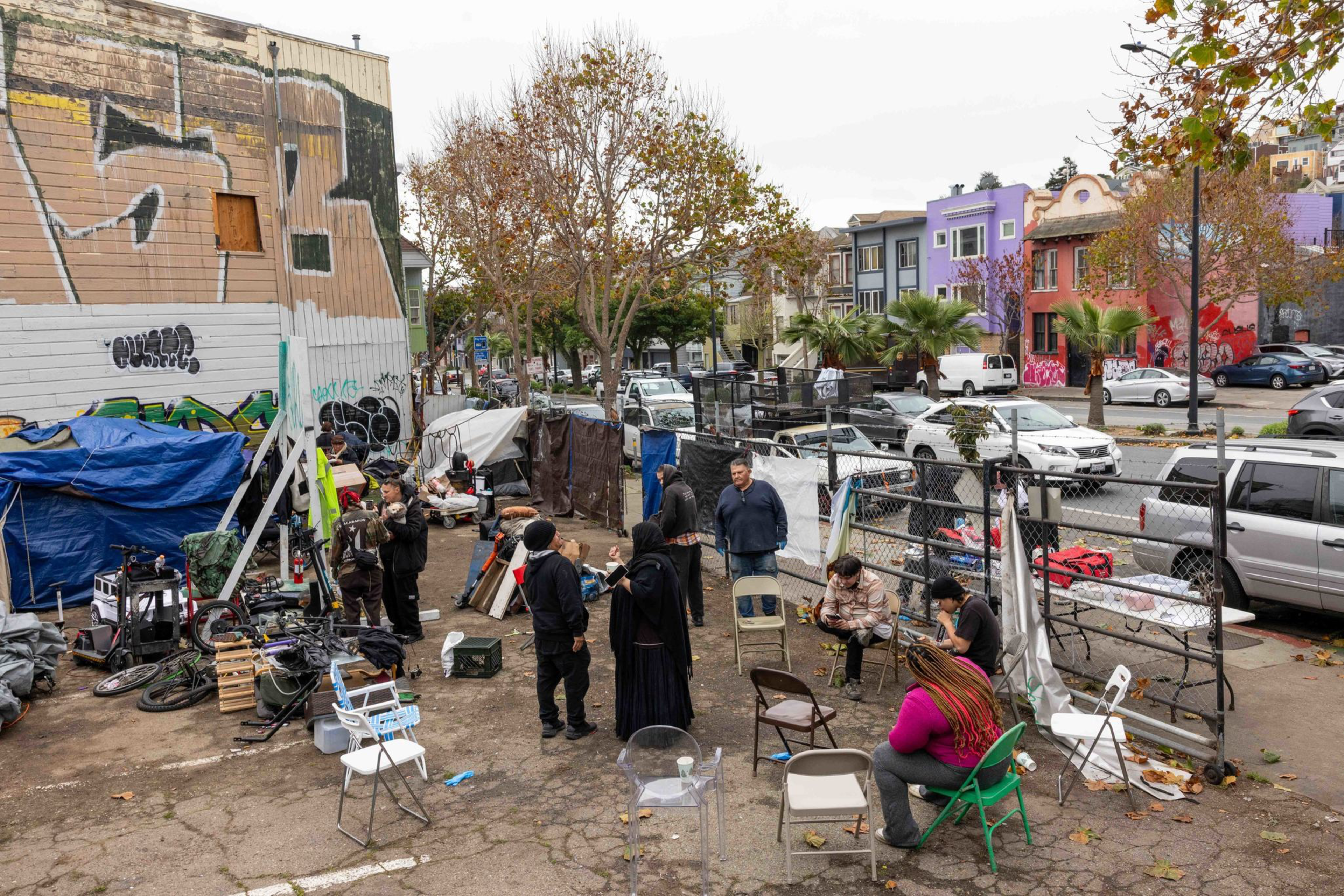 Several people gather and sit on mismatched chairs in a makeshift outdoor area next to a building with graffiti and tents.