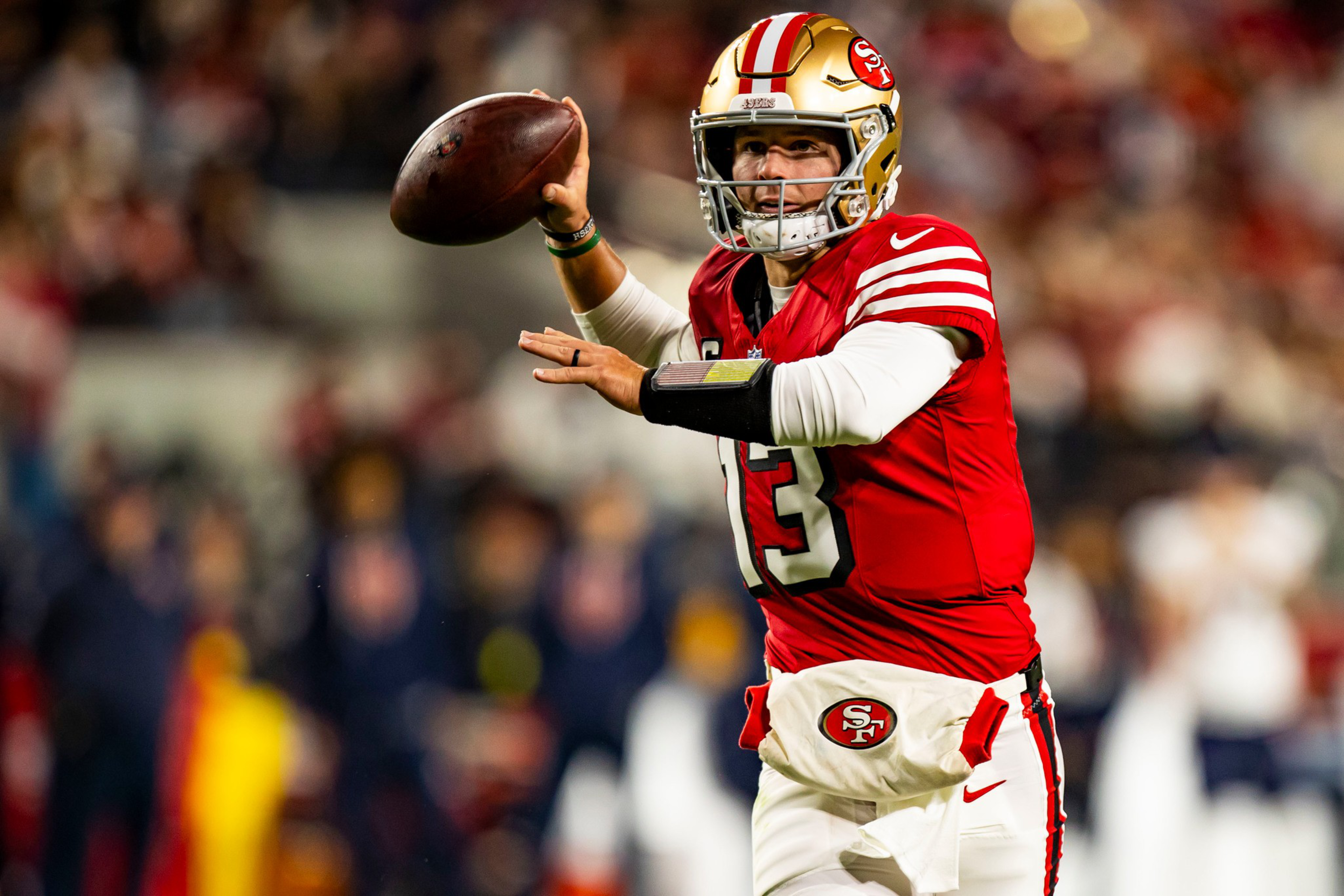 A football player in a red San Francisco 49ers jersey with number 13 is holding a football, preparing to throw it during a game.