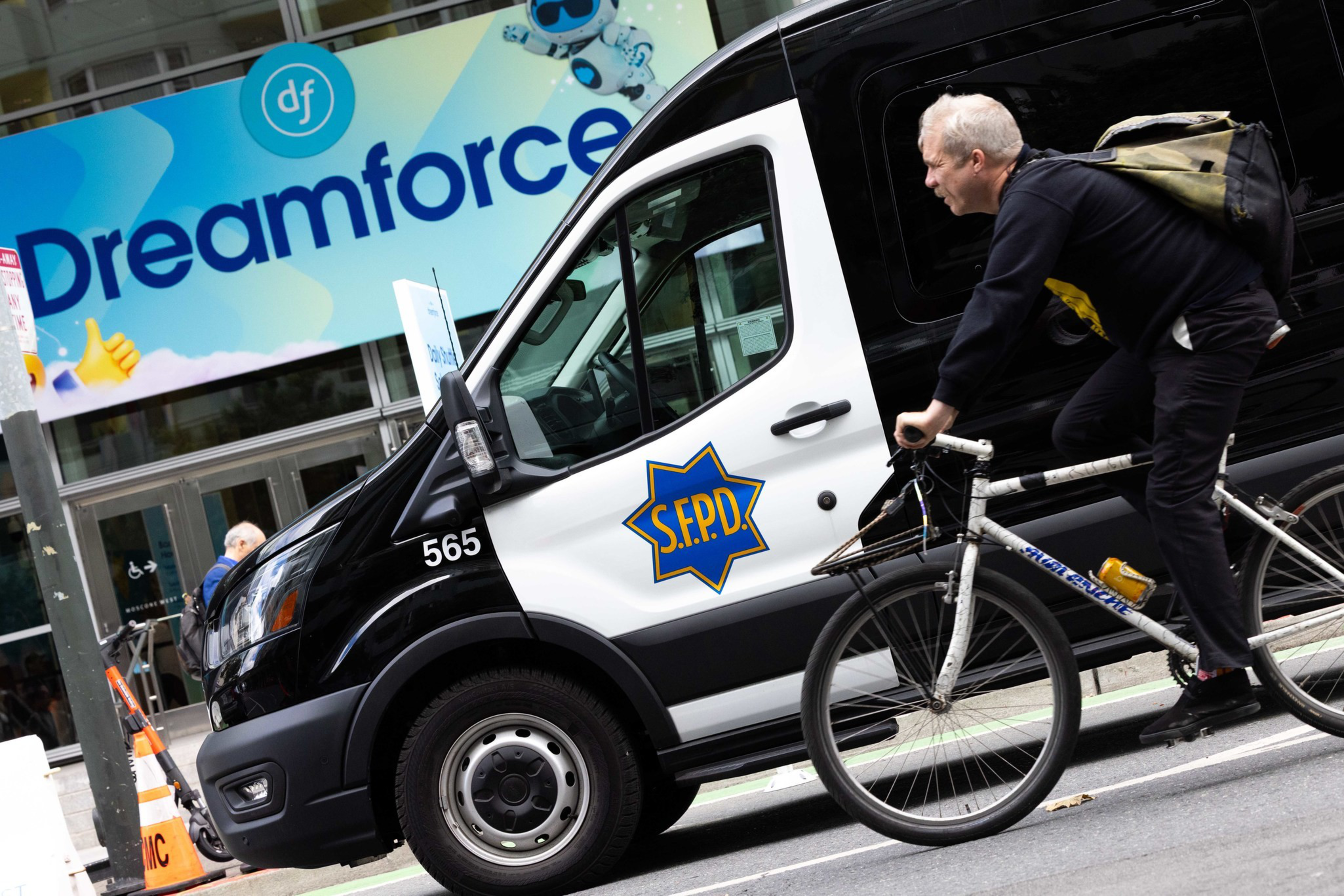 A man with a backpack rides a bicycle past a black-and-white SFPD van parked near a building with a bright “Dreamforce” sign.