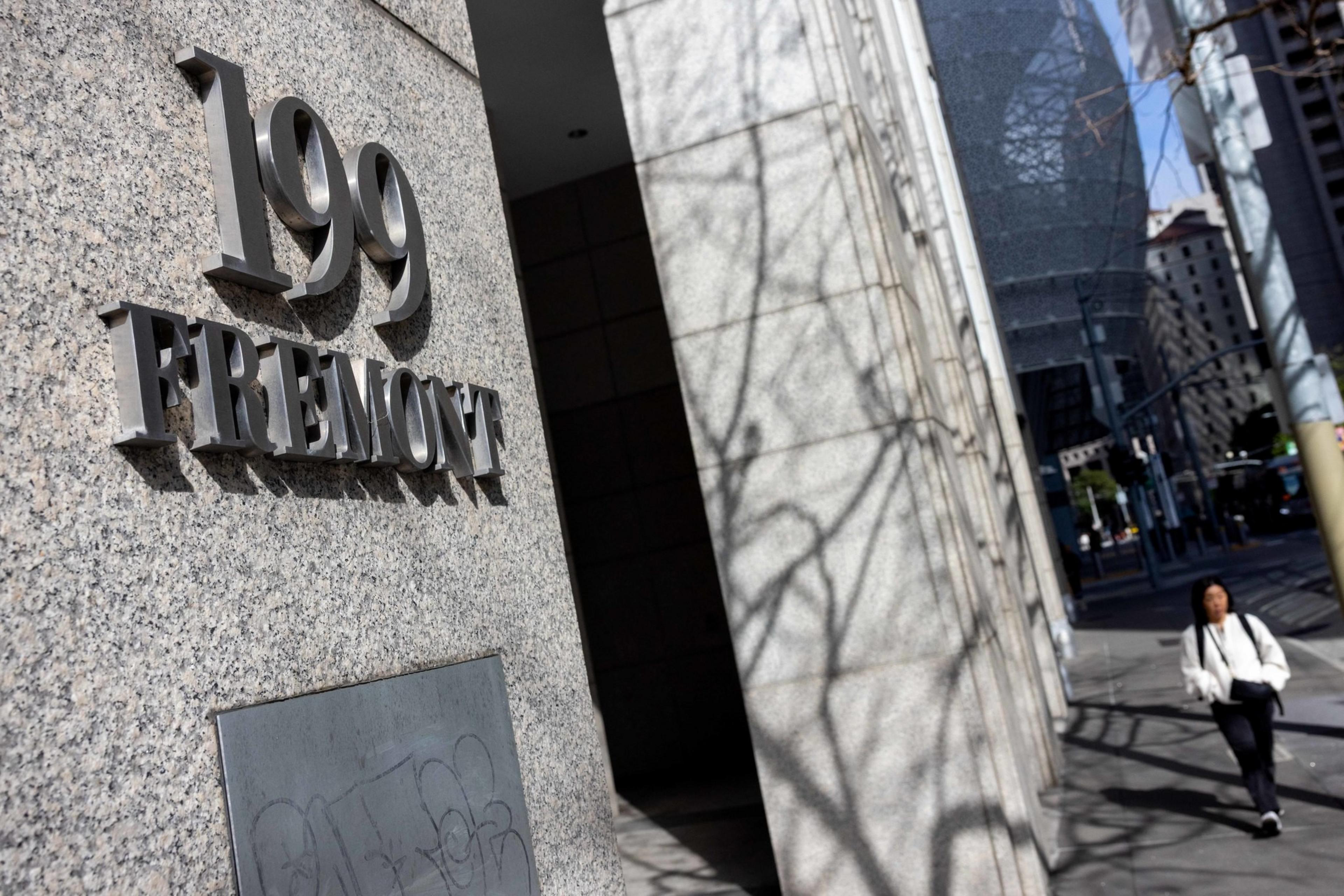 The image shows a building entrance with “199 Fremont” displayed in metal letters on a stone wall, while a person walks on the sidewalk nearby. Shadows of branches are visible on the wall.