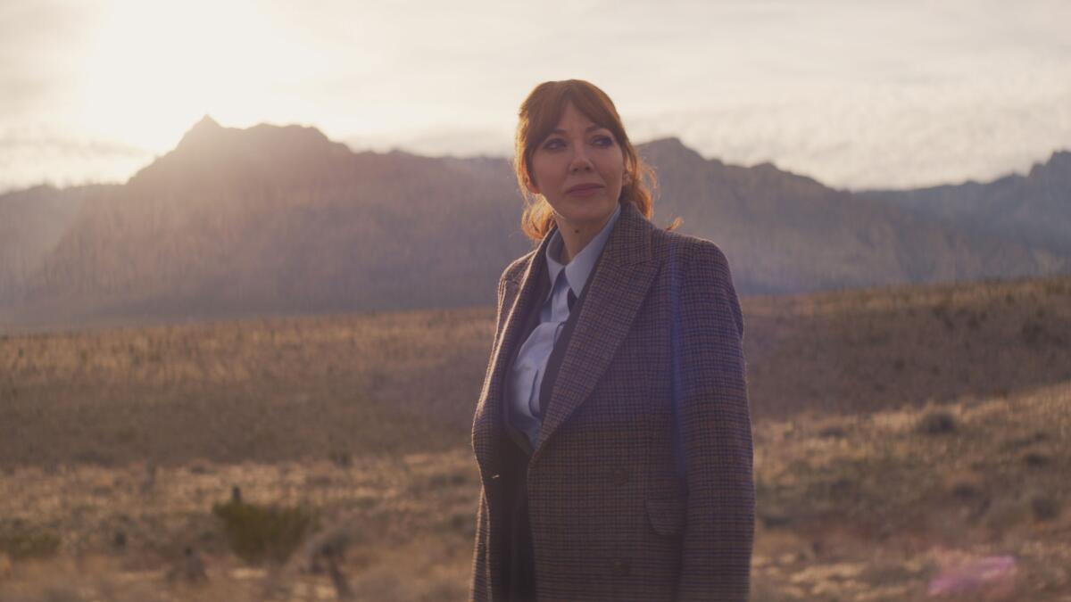 A woman in a blazer, looking to one side, stands outside, mountains in the background
