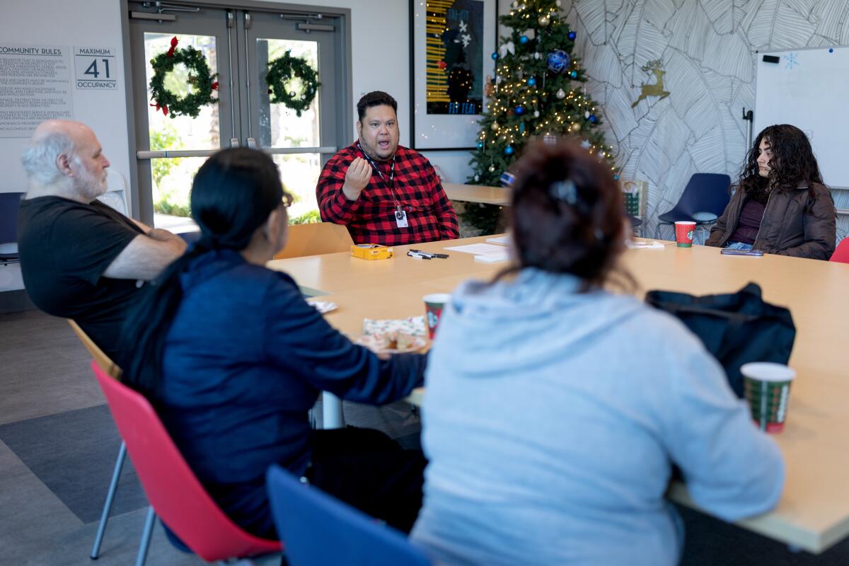 A man with dark hair, in a red plaid shirt, as other people seated around him at a table listen