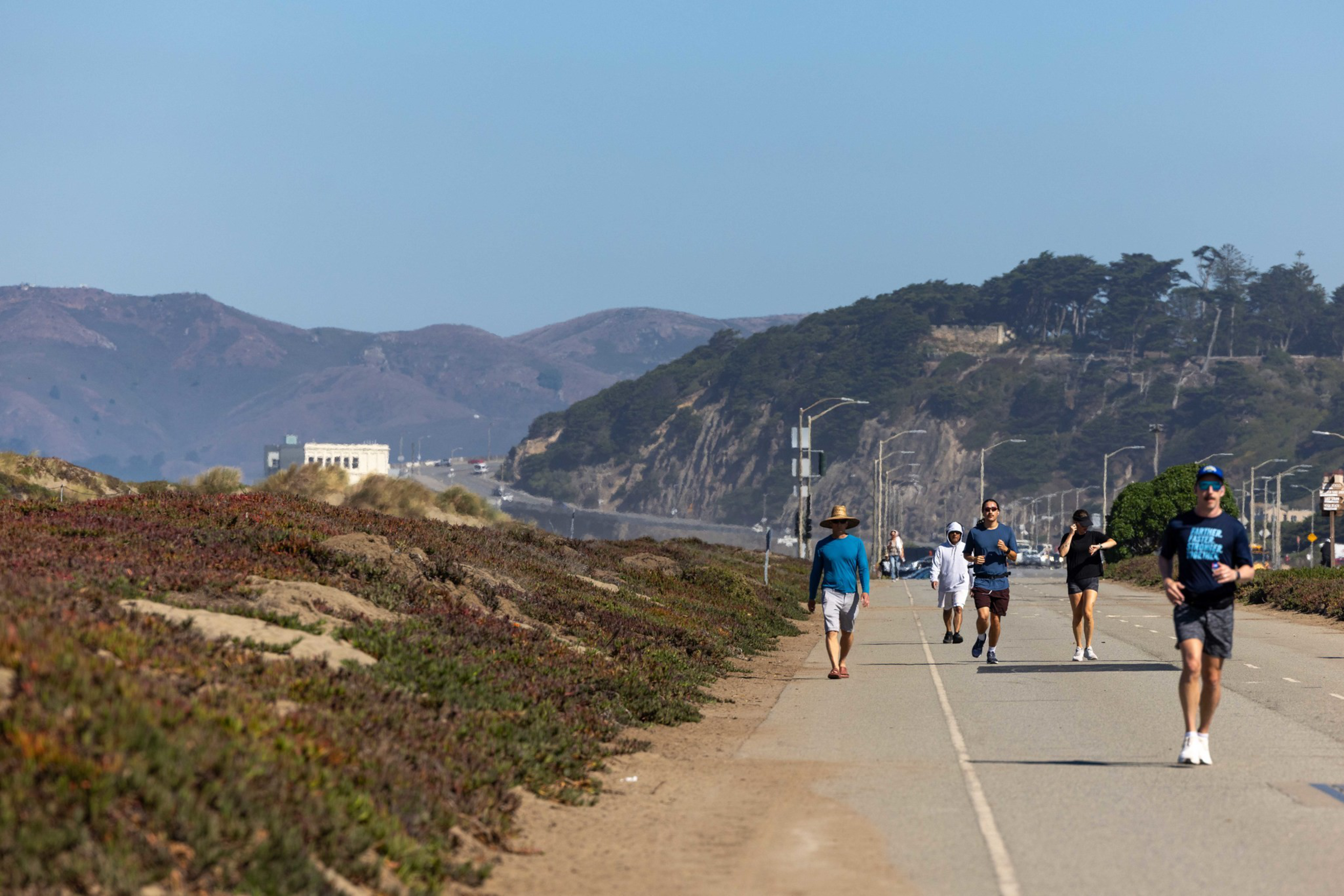 People jog and walk on a paved path along a coastline with hills and a clear blue sky in the background.
