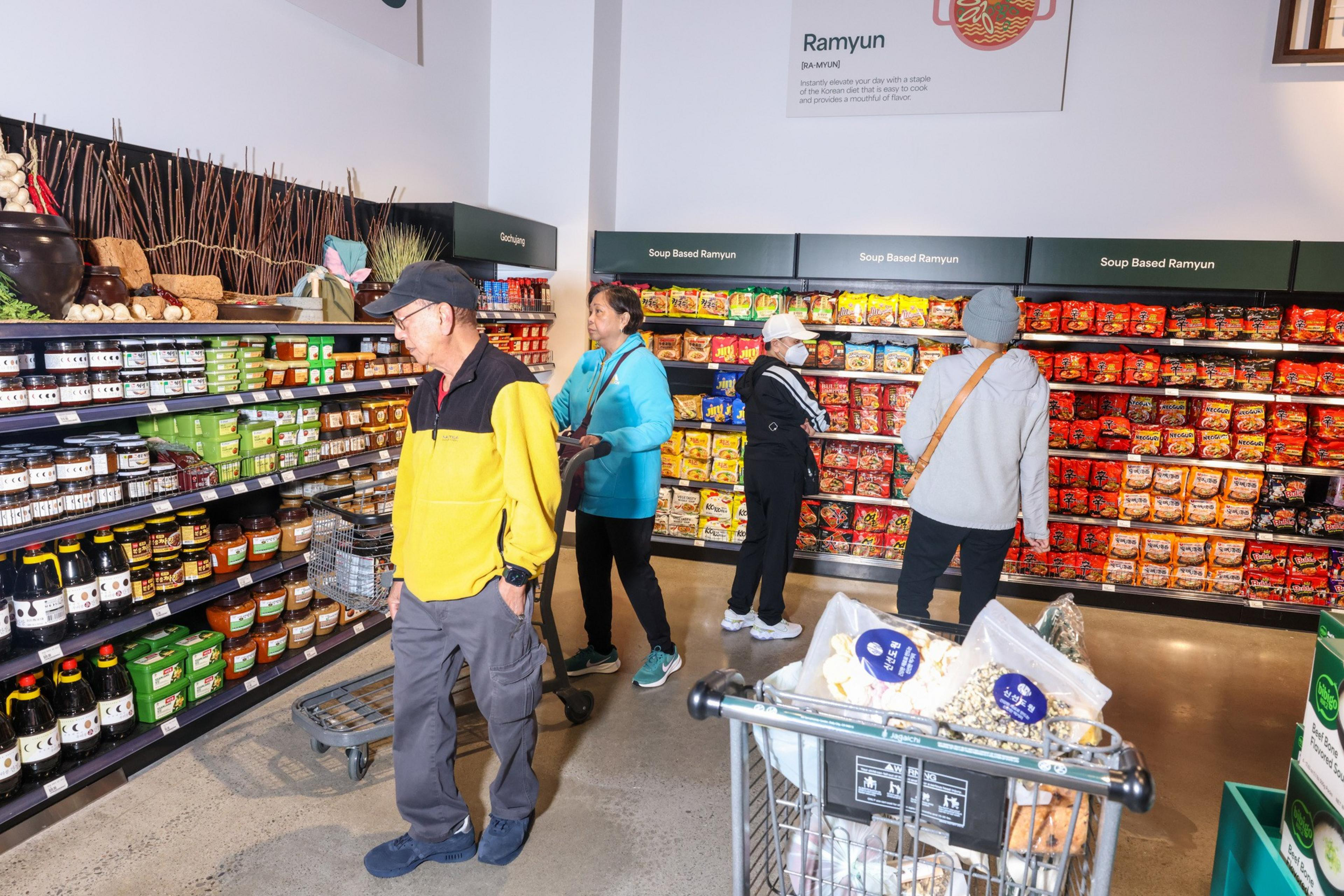 Four people are shopping in a grocery aisle filled with various ramyun and sauces. One person pushes a cart containing packaged goods.