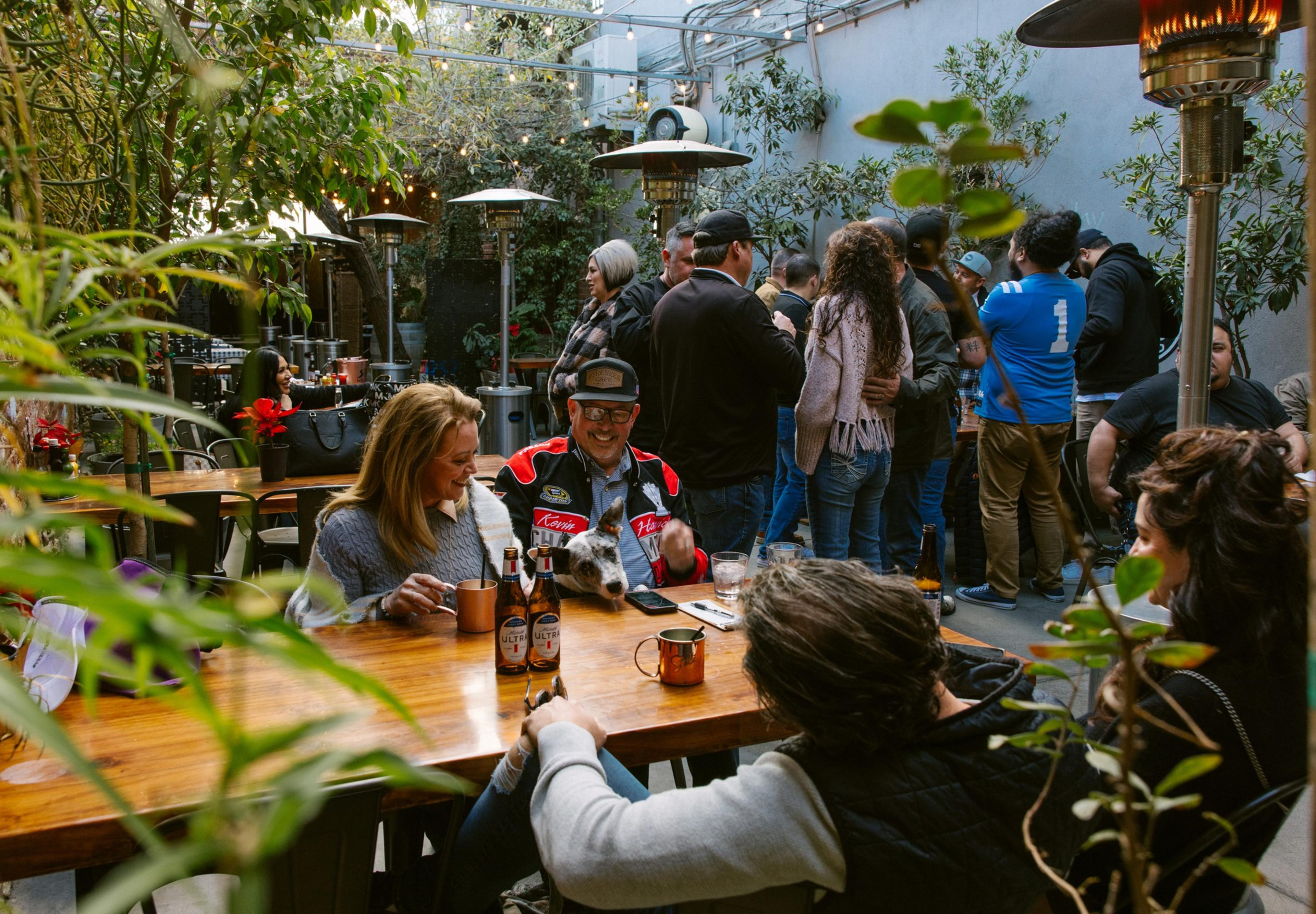 People are gathered at wooden tables in a lively outdoor setting. String lights hang above, and there are heaters. Drinks are on the tables, with a relaxed, social vibe.