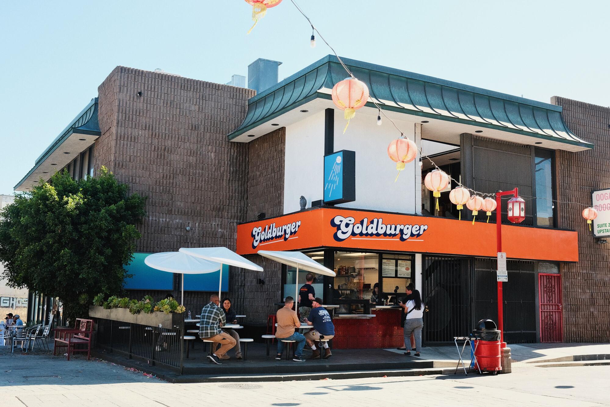 An exterior of Goldburger in Chinatown: guests eat on a patio. Faded red lanterns hang nearby.