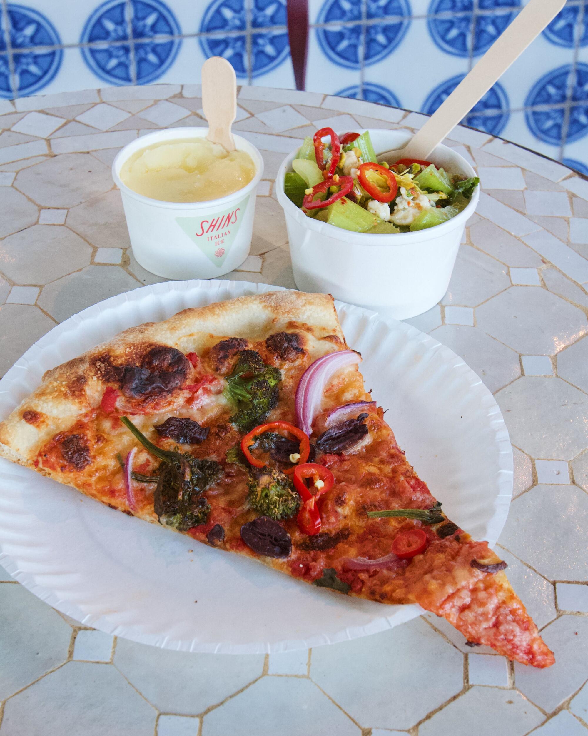 A slice of vegetarian pizza, a cup of celery salad, and an Italian ice on a tiled table at Shins Pizza