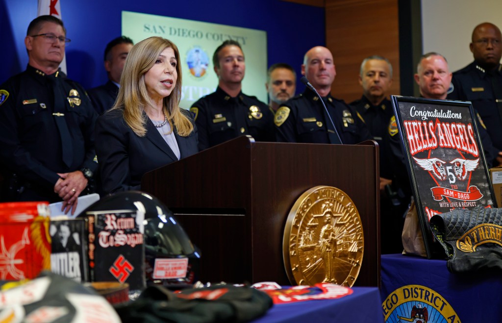 San Diego County District Attorney Summer Stephan announces grand jury indictments while surrounded by police officers and evidence including a "Congratulations Hells Angels" sign, a helmet, and a book with a swastika and the word "Hitler" on it.