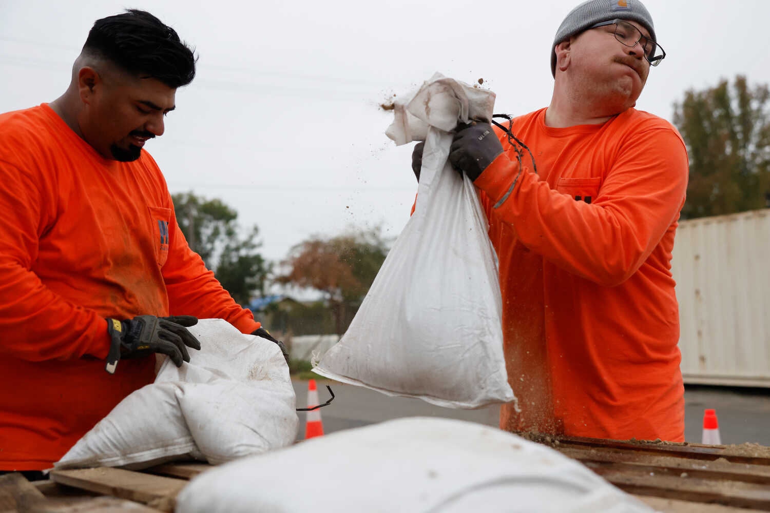 Two men in orange work shirts lift sandbags out of a truck. 
