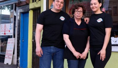 Three people stand together in front of a green storefront with a sign reading "Cocina" and "Mexican Restaurant" on a city street.