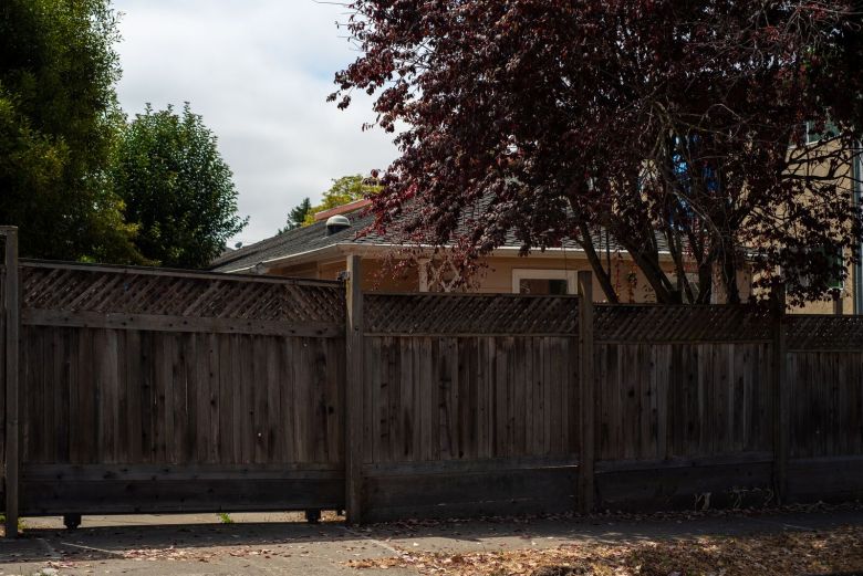 The roofline of a house sticks out from behind a wooden fence along a residential sidewalk