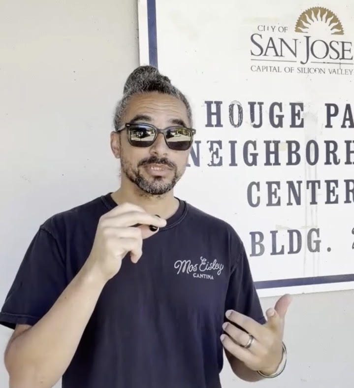 A man in a t-shirt speaking into a small microphone in front of a municipal sign.