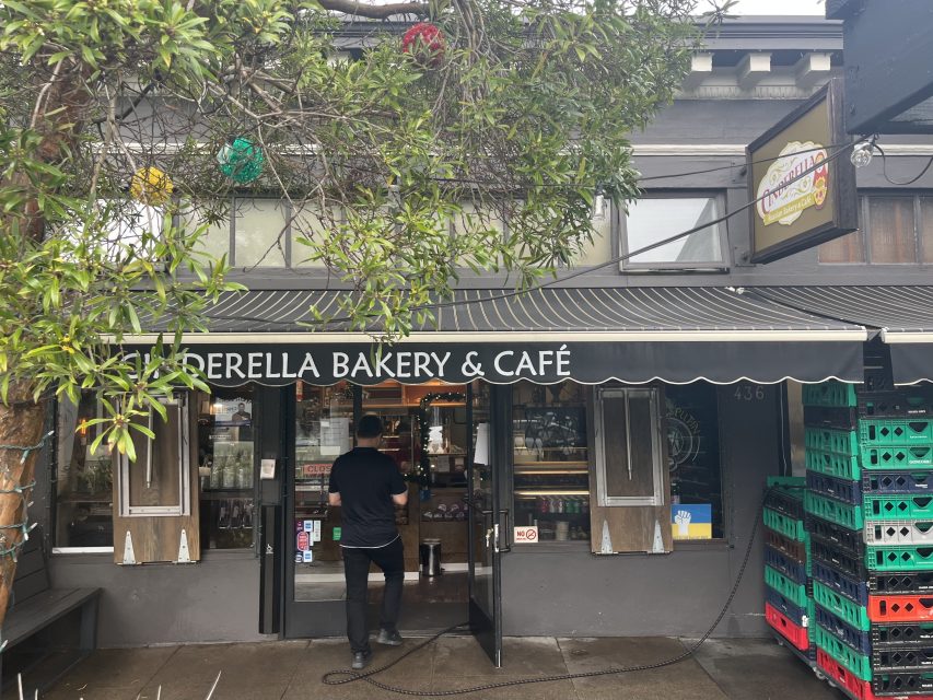 A person enters Perella Bakery & Café, which has a black awning, large front windows, and stacks of plastic bread crates outside.