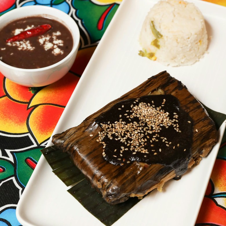 A mole tamal from Guelaguetza sits on a plate with a bowl of white rice and black beans
