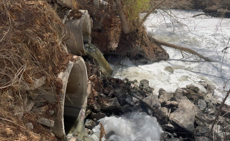 Foam runs through water in sediment around a flowing river with pipes spewing rust colored water into the flow.