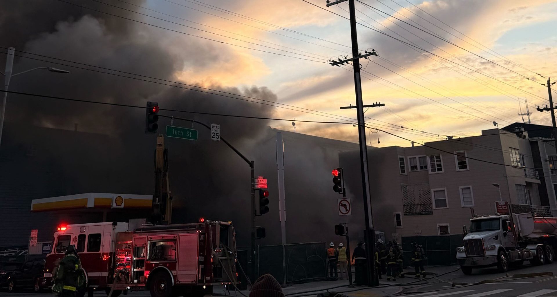 Firefighters respond to a building fire with heavy smoke billowing, fire truck ladders extended, and traffic lights visible at an urban intersection during sunset.