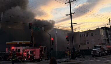 Firefighters respond to a building fire with heavy smoke billowing, fire truck ladders extended, and traffic lights visible at an urban intersection during sunset.