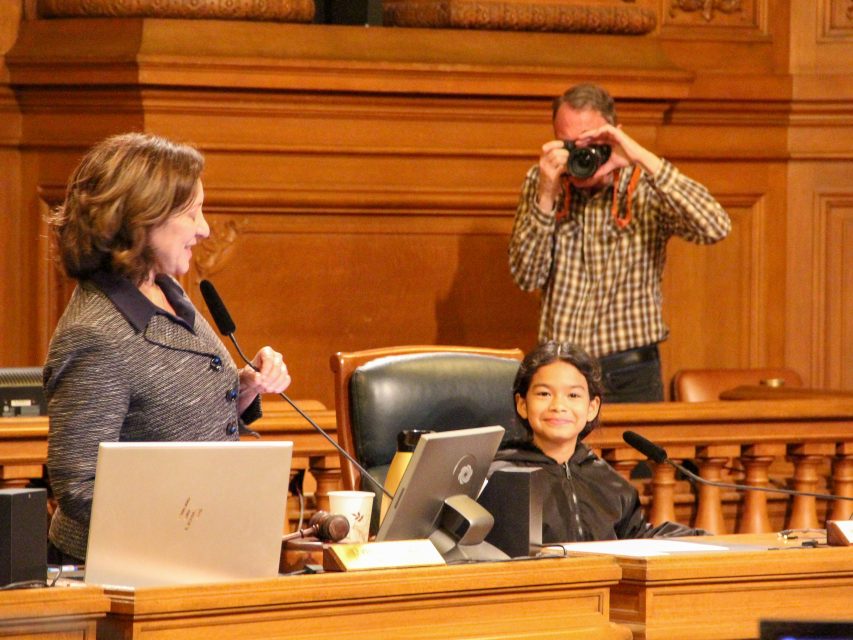 A woman stands at a podium speaking, while a child sits at a large desk and smiles at the camera; a man in the background takes a photo.