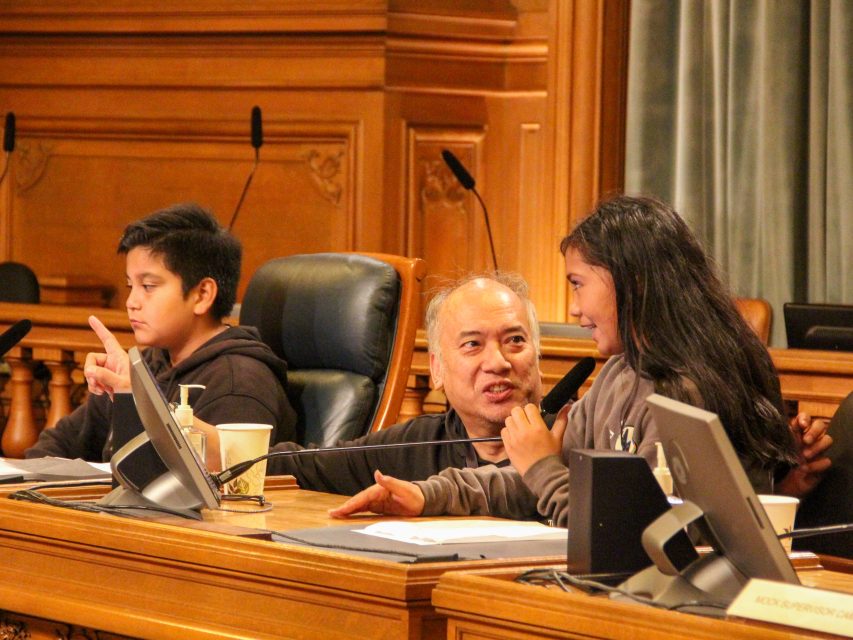 Three people sit at a conference table with microphones; an adult in the center looks at a smiling girl speaking, while a boy sits to the side with his hand raised.