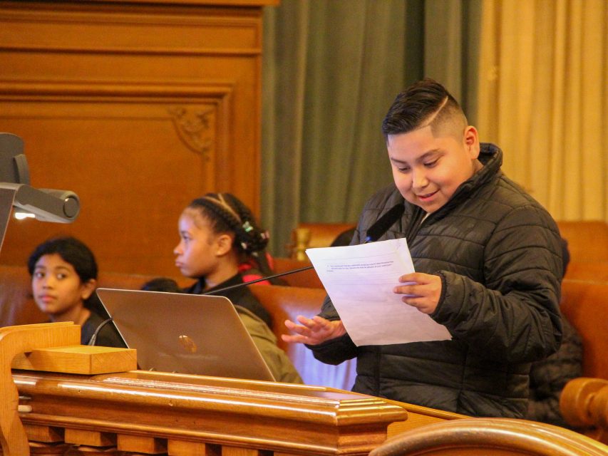 A boy in a black jacket speaks at a podium, holding a sheet of paper, while two other students sit nearby in a formal room.